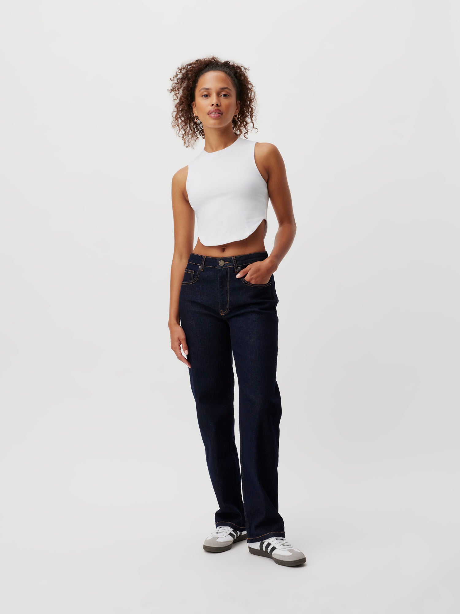 Eine Person mit lockigem Haar steht vor einem weißen Hintergrund, trägt ein ärmelloses weißes Crop Top mit perfekter Passform, dunkelblaue Jeans „Jillian“ von LeGer by Lena Gercke und weiße Sneakers mit schwarzen Streifen. Sie hat eine Hand in der Tasche und einen entspannten Gesichtsausdruck.
