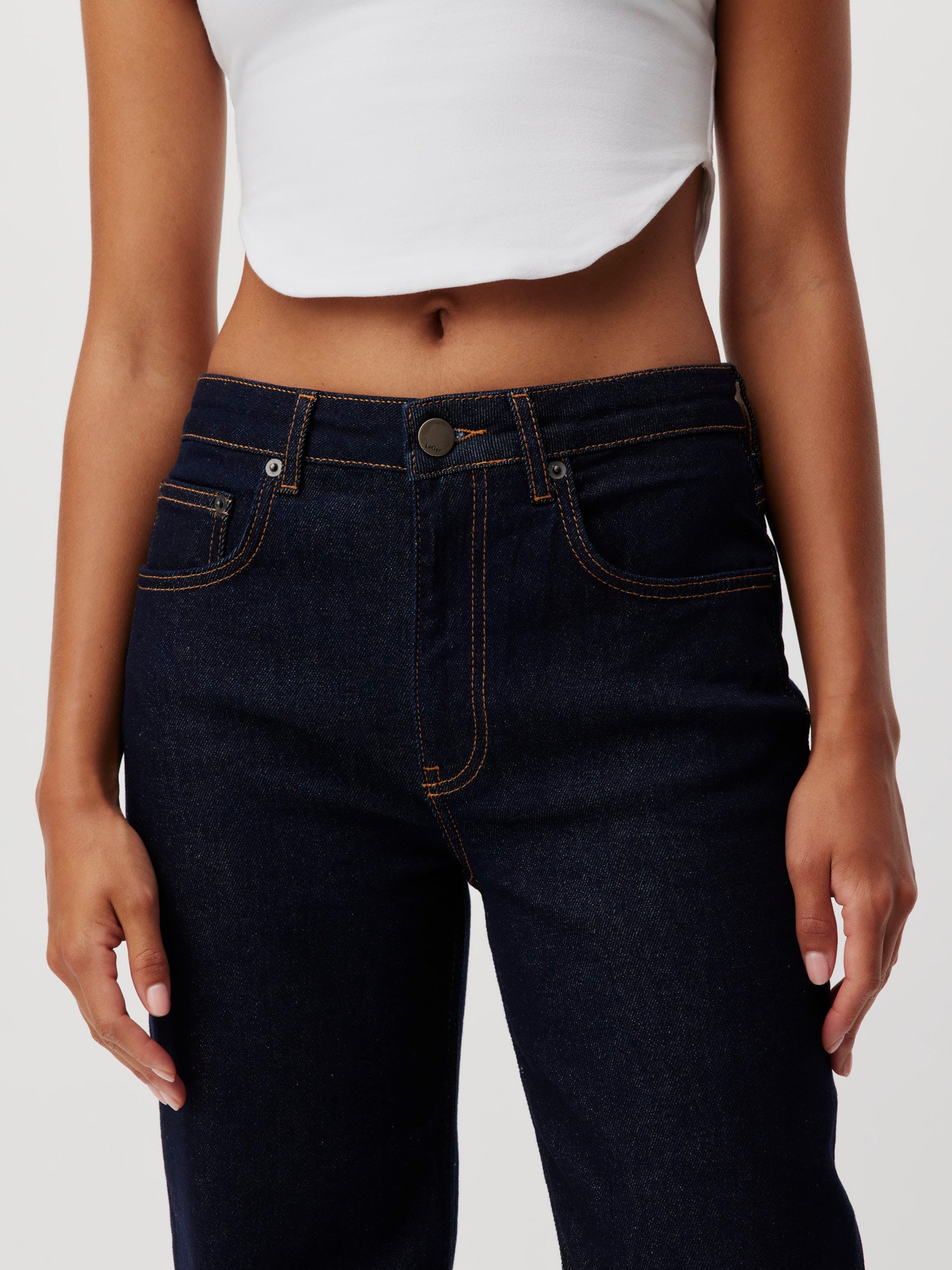 Eine Person in einem weißen Crop Top und dunkler Jeans „Jillian“ von LeGer by Lena Gercke steht mit entspannt herabhängenden Armen da. Der Fokus liegt auf der Körpermitte, die Vorderseite der Jeans mit sichtbaren Nähten und dunklem Knopfverschluss wird freigelegt und betont die tadellose Passform. Der Hintergrund ist schlicht weiß.