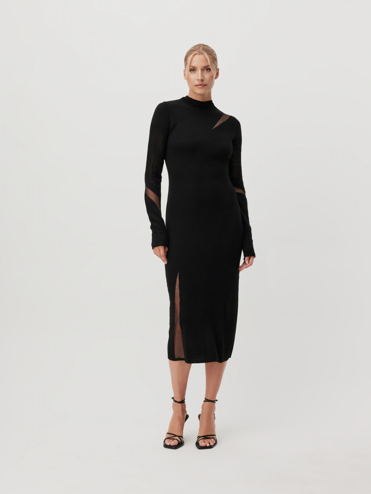 Eine Frau, die das Kleid „Lenio“ von LeGer by Lena Gercke trägt – ein elegantes schwarzes Midikleid mit durchsichtigen Mesh-Ausschnitten und langen Ärmeln –, steht vor einem schlichten weißen Hintergrund und trägt dazu schwarze Pumps.