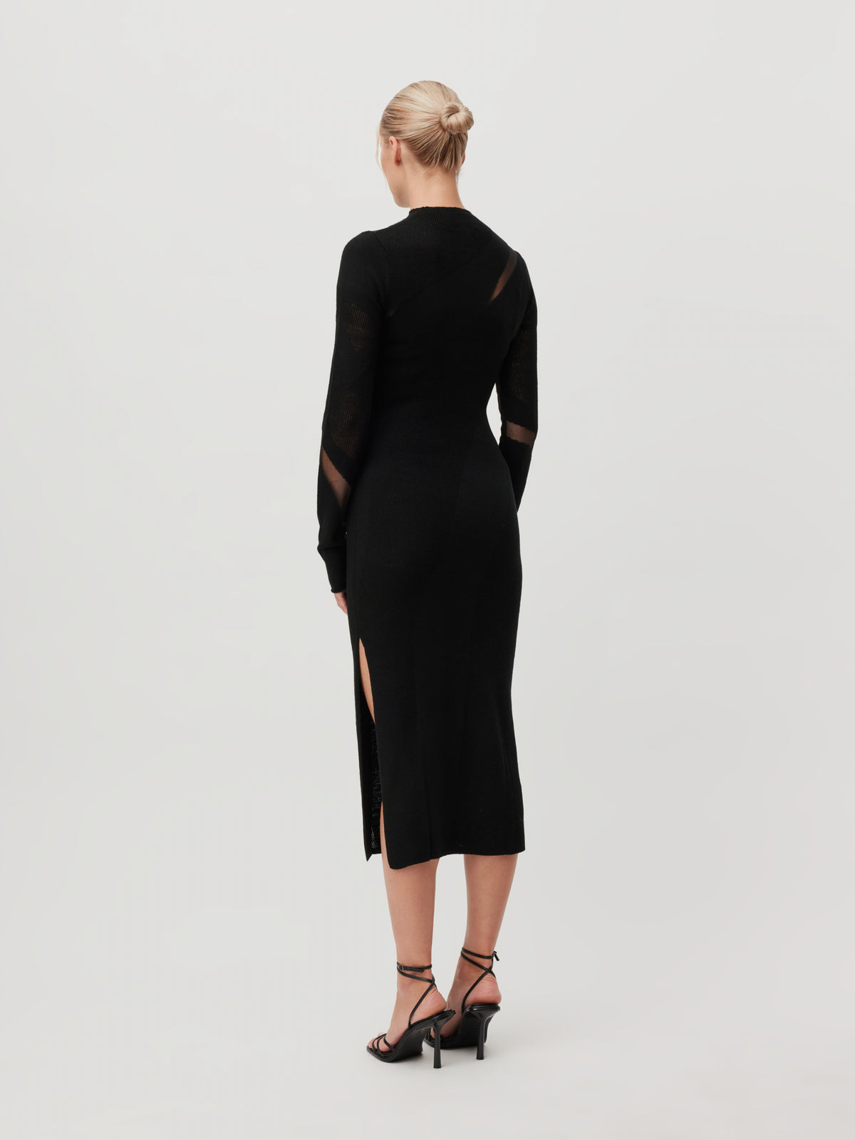 Eine Frau mit blonden Haaren, die zu einem Dutt hochgesteckt sind, trägt das schwarze Midikleid „Lenio“ von LeGer by Lena Gercke mit 3/4-Ärmeln und Schlitz sowie schwarze Sandalen mit Absatz. Sie steht vor einem schlichten, hellen Hintergrund und ist von der Kamera abgewandt.