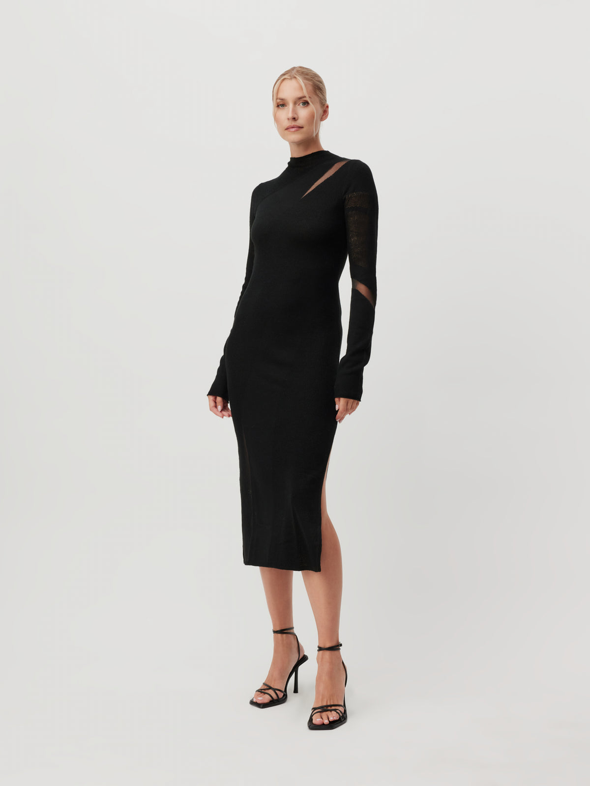 Eine Frau steht vor einem schlichten weißen Hintergrund und trägt das Kleid „Lenio“ von LeGer by Lena Gercke – ein elegantes schwarzes Midikleid mit langen Ärmeln, einem Seitenschlitz und Ausschnittdetails –, dazu schwarze Sandalen mit Absatz.