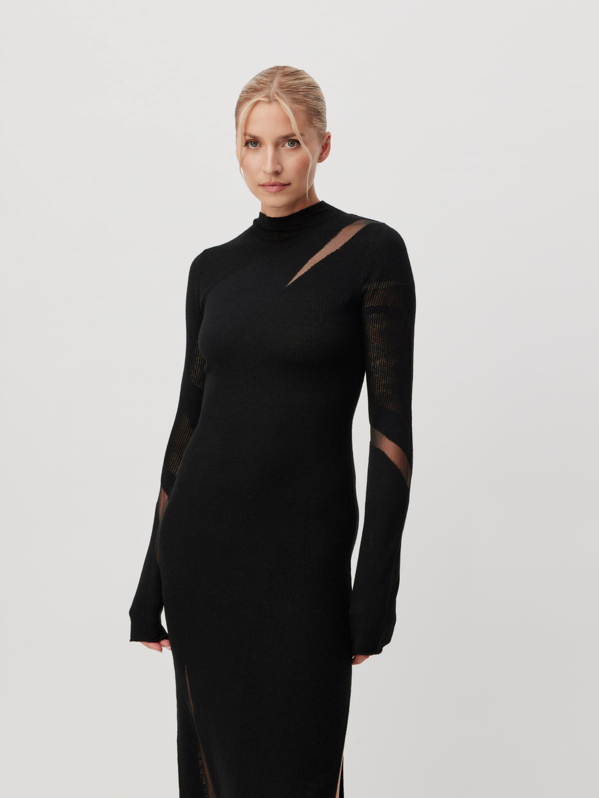 Eine Frau mit blonden Haaren, die zu einem tief sitzenden Dutt gesteckt sind, trägt das Kleid „Lenio“ von LeGer by Lena Gercke – ein elegantes, schwarzes, eng anliegendes Langarm-Modell mit transparenten Ausschnittdetails – und steht vor einem schlichten, hellen Hintergrund.