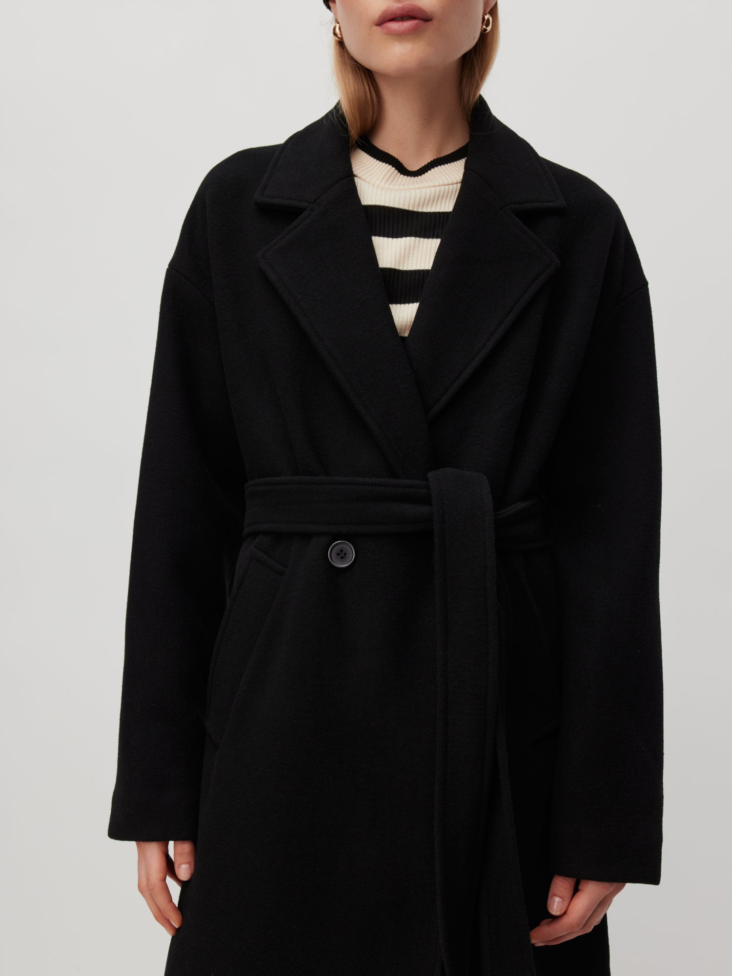 Eine Frau im Dena Coat von LeGer by Lena Gercke, mit Gürtel und normaler Länge, steht vor einem schlichten Hintergrund. Unter dem Mantel trägt sie ein schwarz-weiß gestreiftes Oberteil, das eine elegante Passform betont. Ihr schulterlanges Haar unterstreicht ihren Look, ergänzt durch kleine Creolen.
