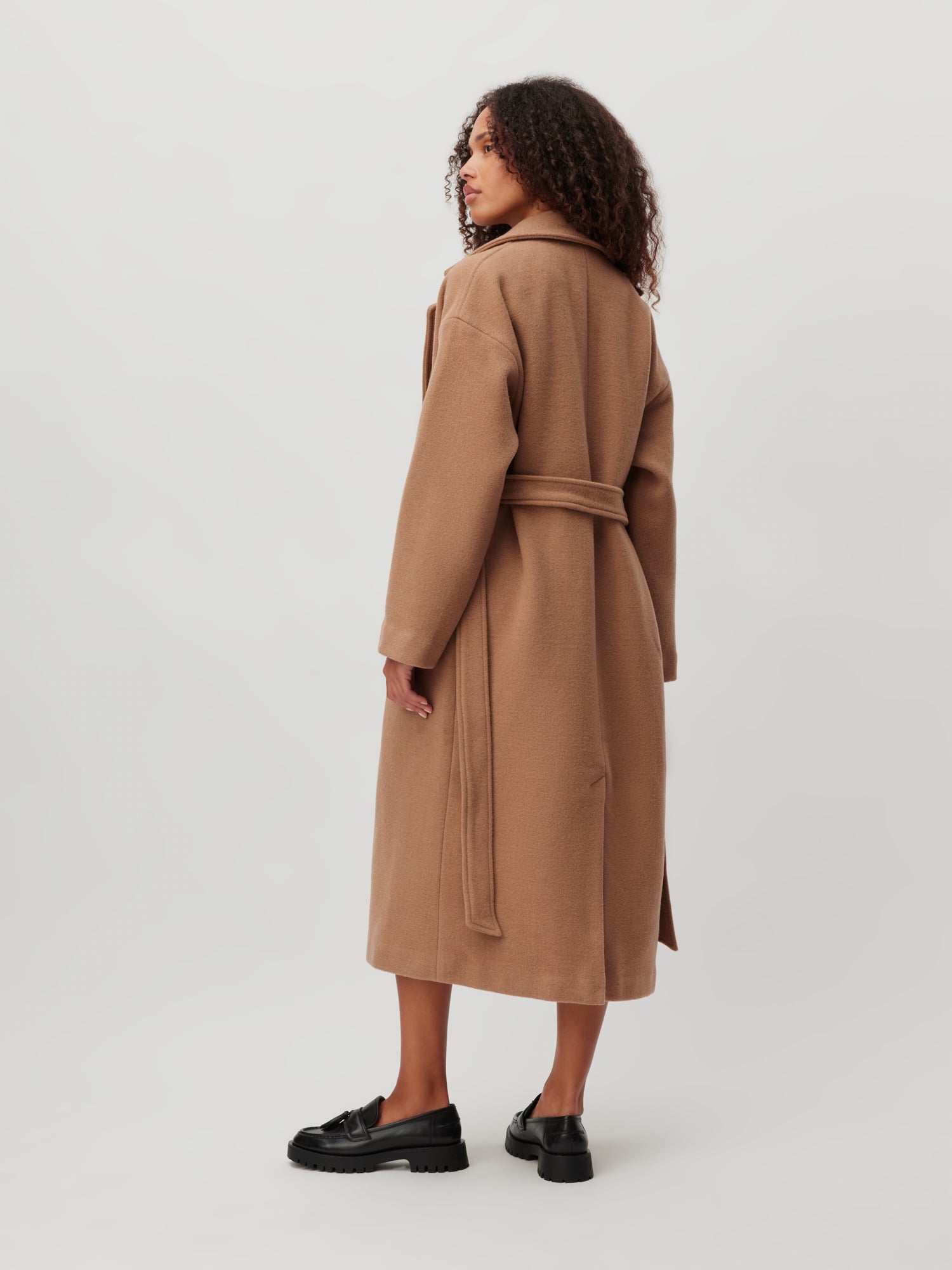 Vor einem schlichten Hintergrund steht eine Person mit lockigem Haar, die den Dena Coat von LeGer by Lena Gercke trägt. Der lange, kamelfarbene Mantel hat eine normale Passform und einen Gürtel in der Taille, der das Ensemble perfekt ergänzt, während sie in schwarzen Slippern zur Seite blickt.