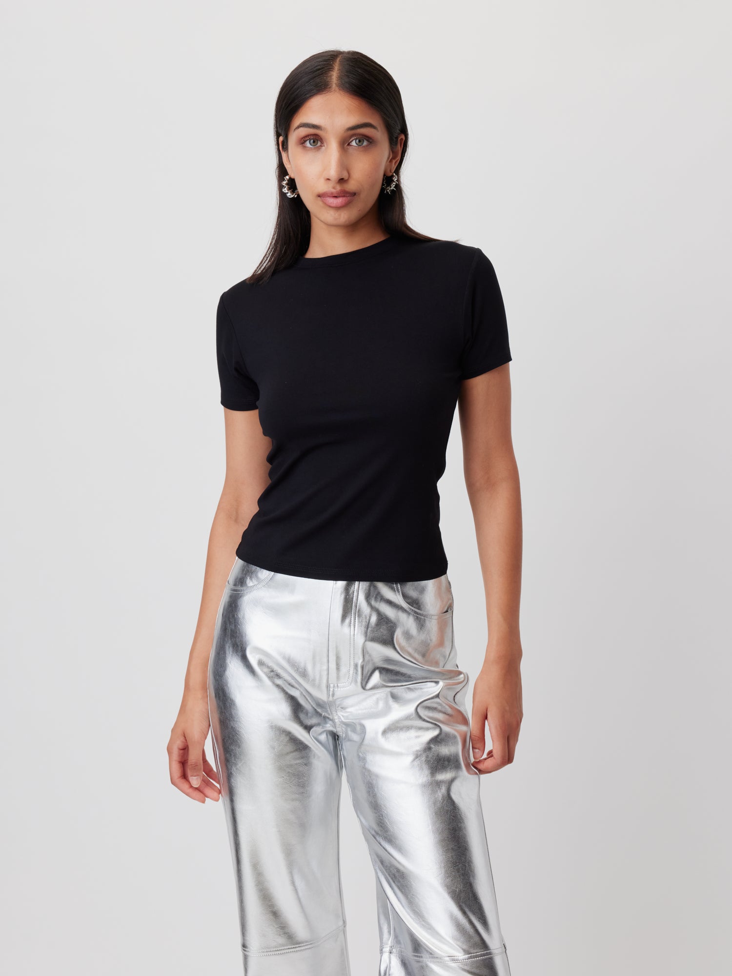 Lena Gercke modelliert das LeGer by Lena Gercke T-Shirt 'Doreen'. Sie stylt das taillierte schwarze Oberteil mit einer silbernen Metallic-Hose, während sie selbstbewusst vor einem schlichten, hellen Hintergrund steht und direkt in die Kamera schaut.