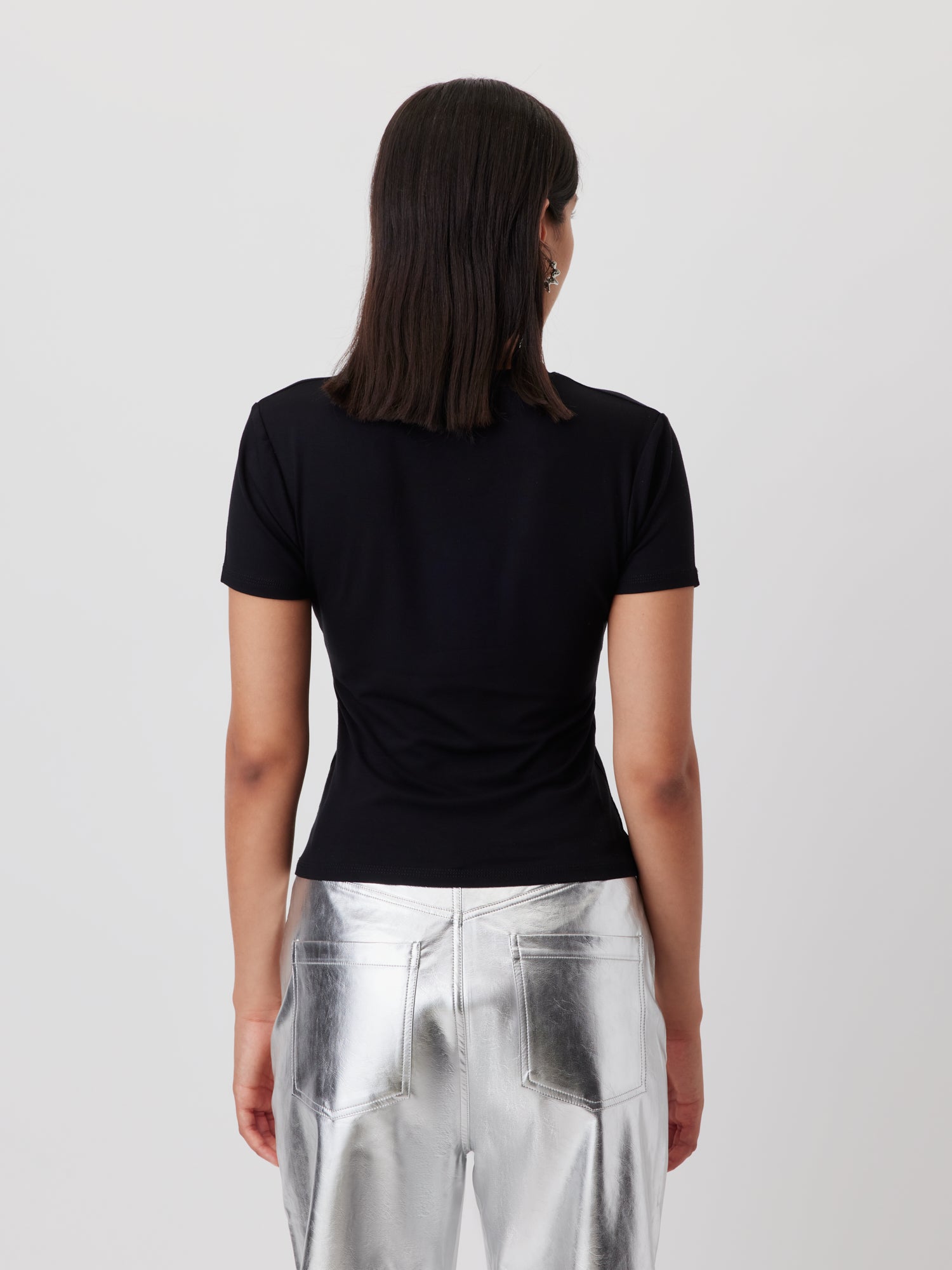 Eine Person mit glattem, schulterlangem dunklem Haar ist von hinten zu sehen. Sie trägt das T-Shirt "Doreen" von LeGer by Lena Gercke und eine silbern glänzende Metallhose vor einem schlichten, hellen Hintergrund.