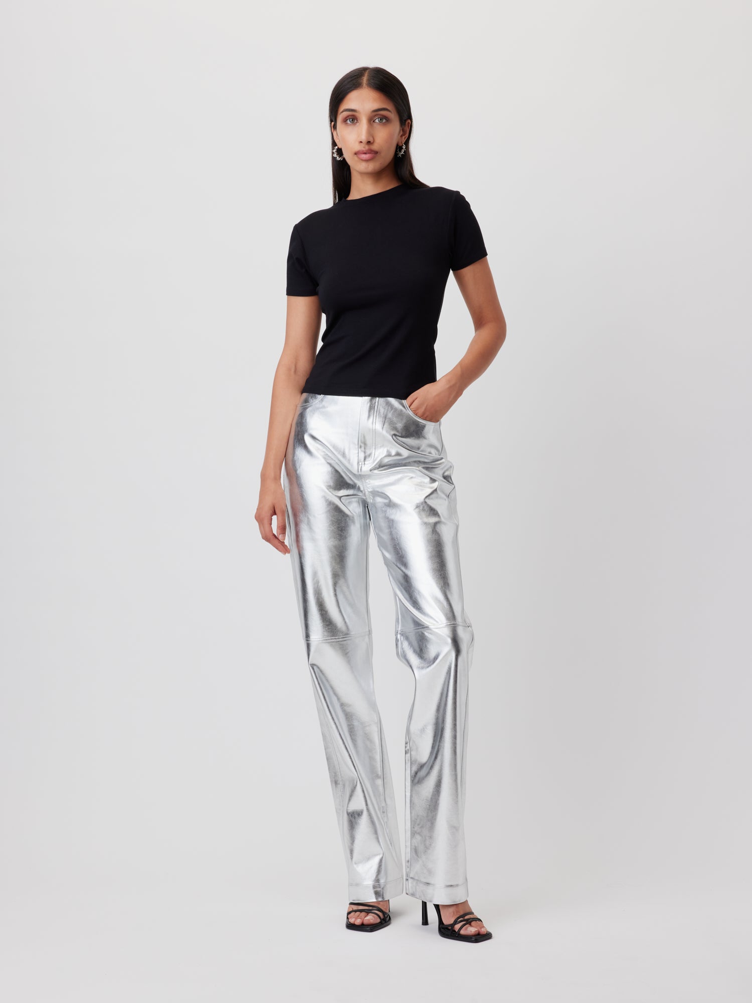 Eine Frau mit langen dunklen Haaren trägt das schwarze T-Shirt 'Doreen' von LeGer by Lena Gercke, eine silbern glänzende Metallic-Hose mit geradem Bein und schwarze Sandalen mit Absatz vor einem weißen Hintergrund. Sie posiert mit einer Hand in der Tasche im Stil von Lena Gercke.