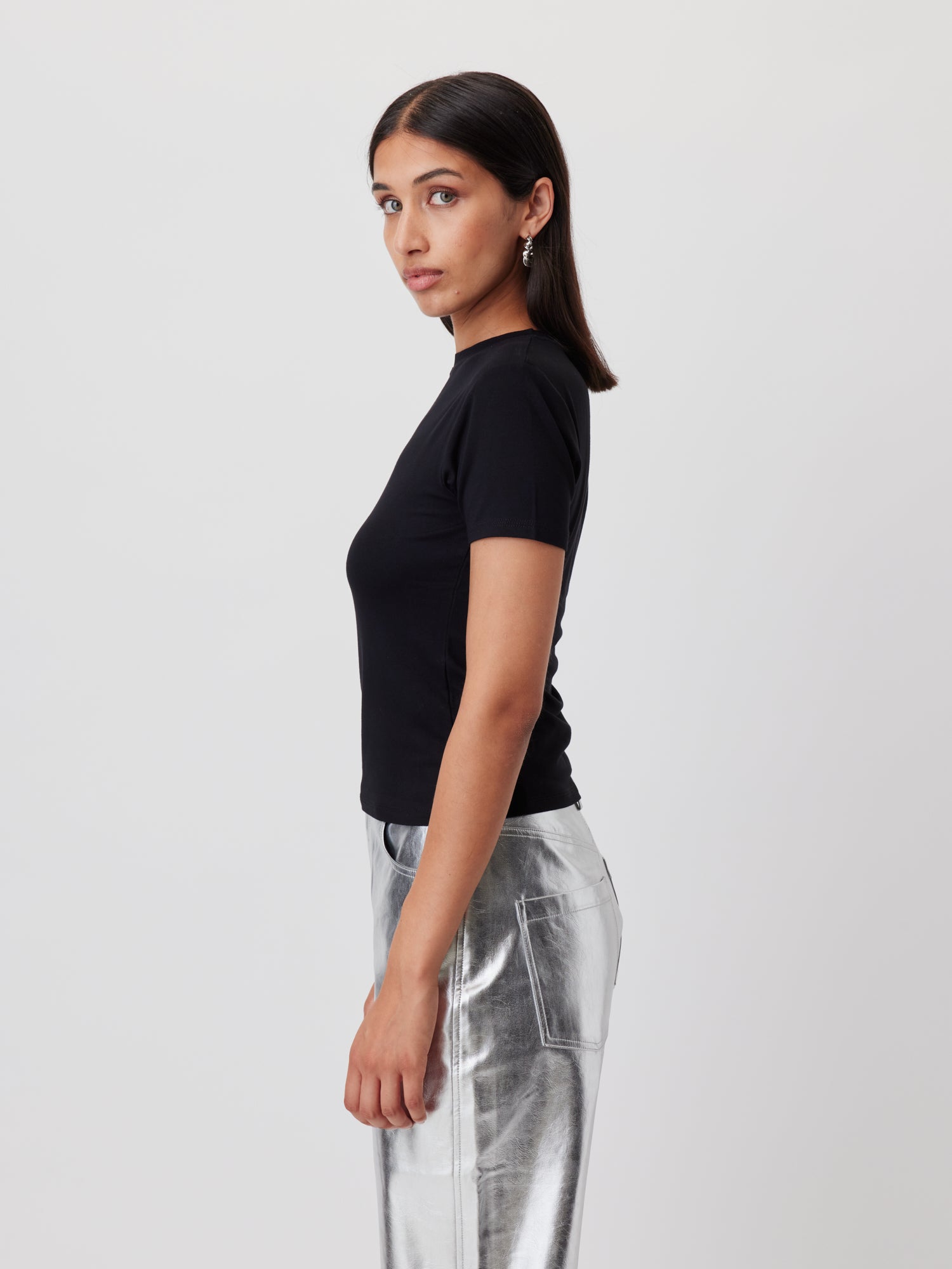 Frau mit glattem dunklem Haar trägt ein tailliertes LeGer by Lena Gercke T-Shirt 'Doreen' und eine silbern glänzende Metallic-Hose und steht im Profil vor einem schlichten hellen Hintergrund.