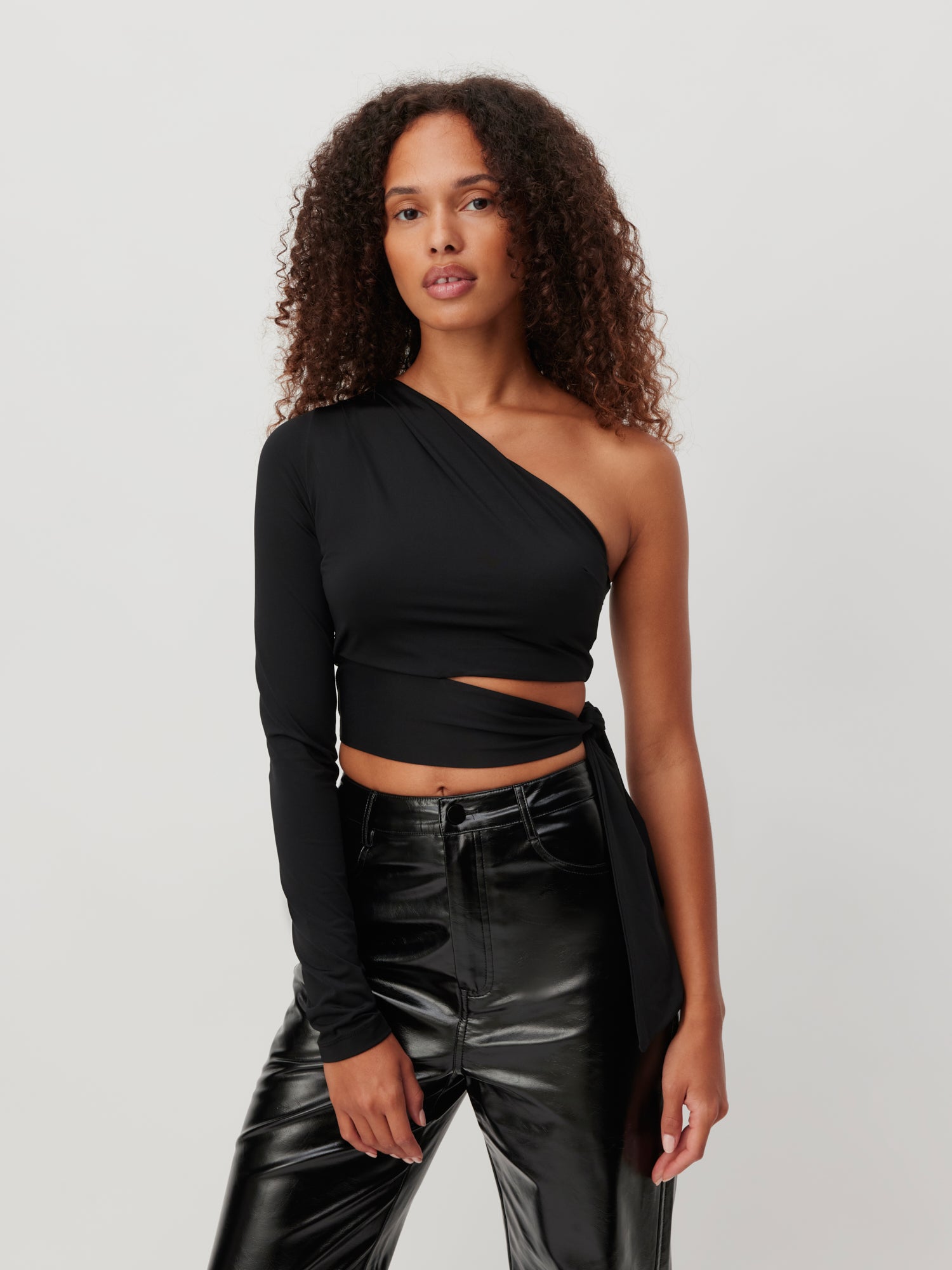 Eine Person mit lockigem Haar trägt ein stylisches, asymmetrisches schwarzes Top namens „Shirt ‚Leia‘“ von LeGer by Lena Gercke, das einen Langarm und ein Cut-Out-Design aufweist. Dazu hat sie eine hoch taillierte, glänzende schwarze Hose in Größe 36 kombiniert und steht selbstbewusst vor einem schlichten weißen Hintergrund.