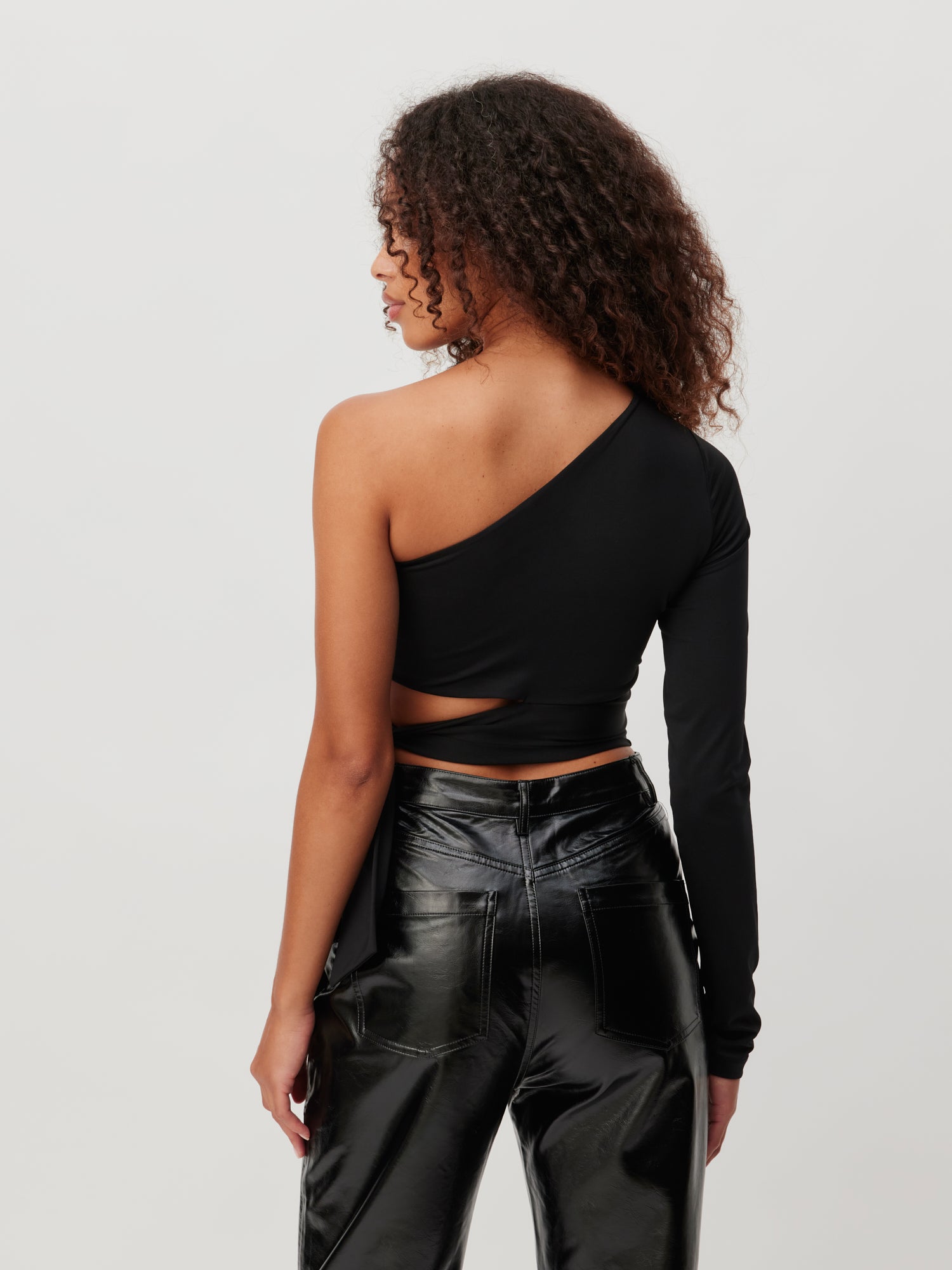 Eine Person mit lockigem Haar steht mit dem Rücken zur Kamera und trägt ein schwarzes One-Shoulder-Top, Größe 36, von Shirt 'Leia' von LeGer by Lena Gercke. Das Top hat an einem Arm einen langen Ärmel und auf der anderen Seite ein Cut-out-Design. Dazu trägt sie eine glänzende schwarze Hose.
