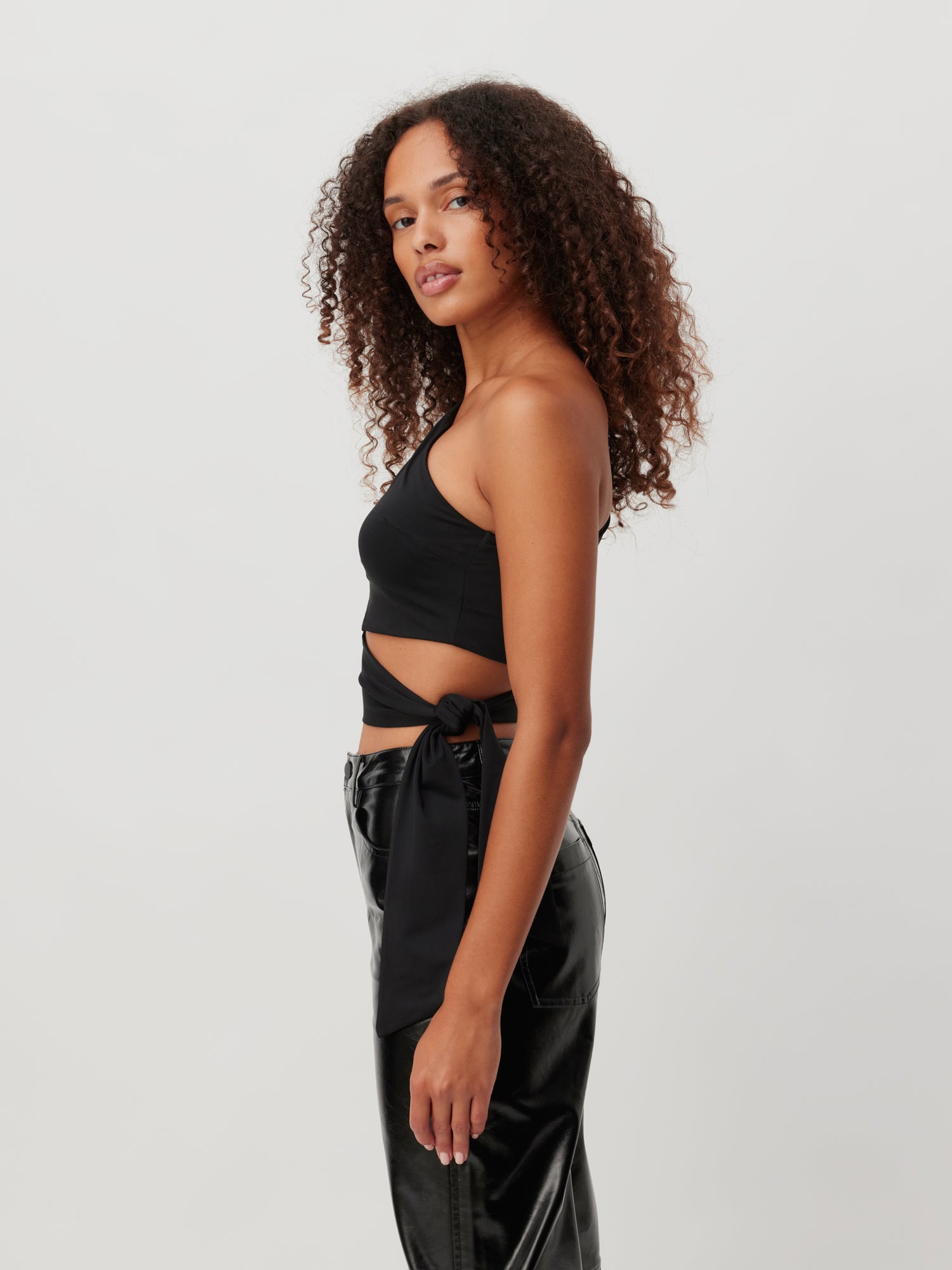 Eine Frau mit lockigem Haar steht im Profil und trägt ein schwarzes, einschultriges Crop-Top namens „Shirt ‚Leia‘“ von LeGer by Lena Gercke und eine schwarze Lederhose, Größe 36. Vor einem schlichten, hellen Hintergrund blickt sie mit neutralem Gesichtsausdruck in die Kamera.