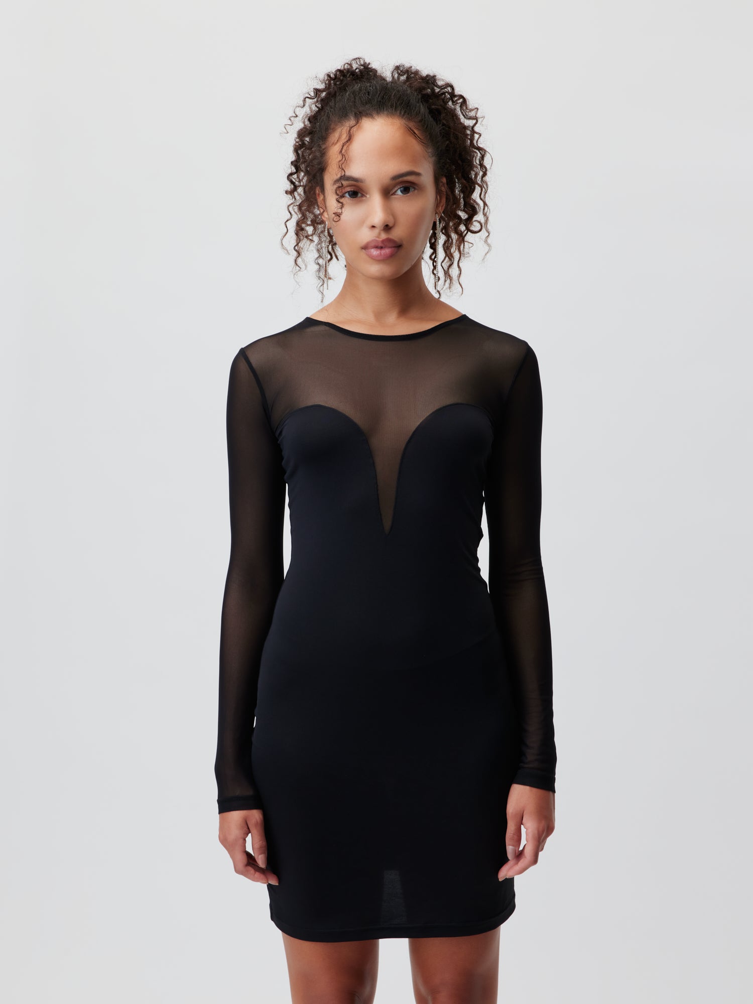 Eine Person steht vor einem schlichten weißen Hintergrund und trägt das „Antonietta Dress“ von LeGer by Lena Gercke. Dieses elegante schwarze Kleid hat durchsichtige lange Ärmel, einen halbtransparenten Ausschnitt und ein herzförmiges Design, wodurch es auffällt. Die lockigen Haare der Person sind ordentlich zurückgebunden.