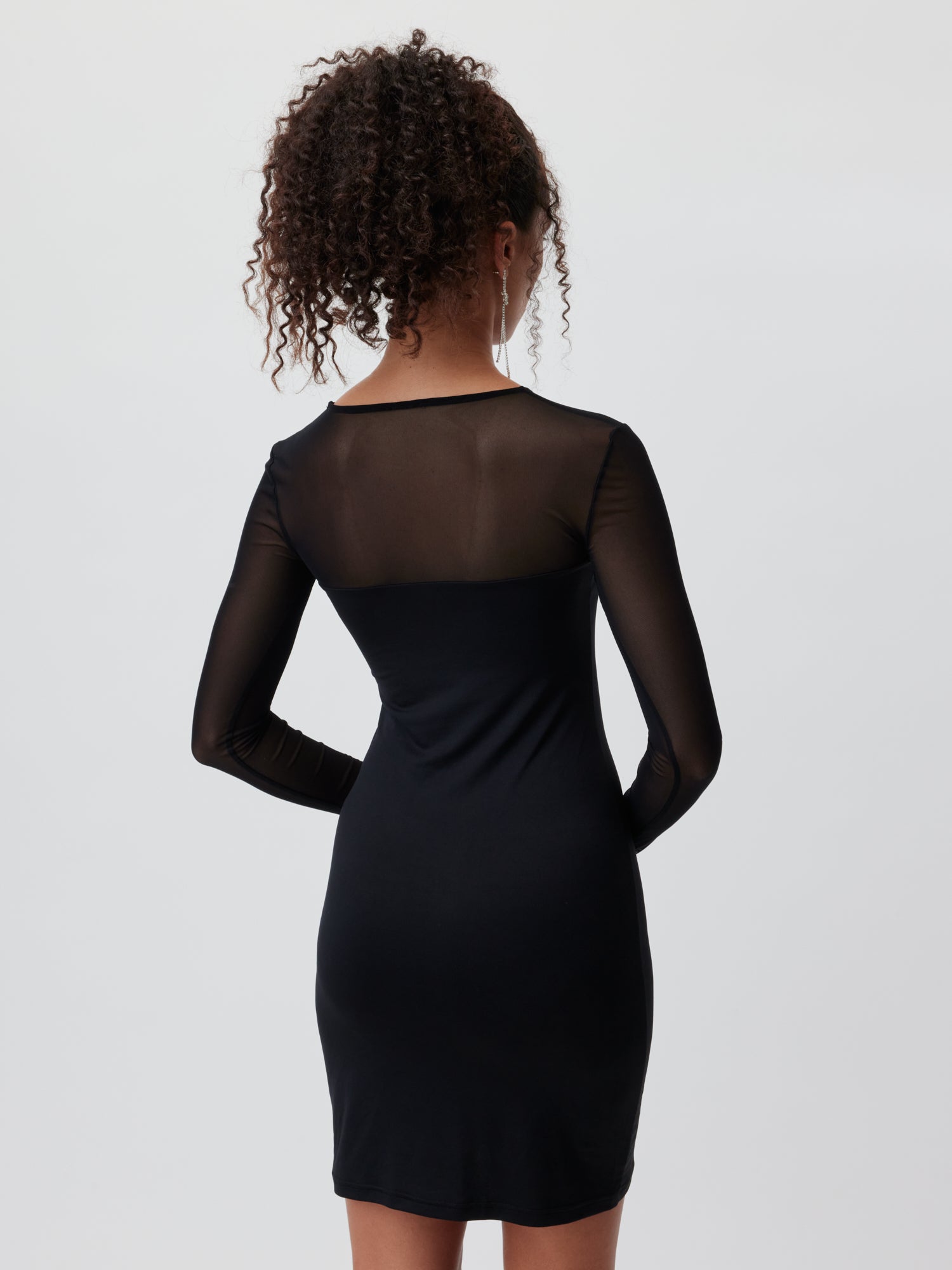 Eine Person mit lockigem Haar trägt ein schwarzes Antonietta-Kleid von LeGer by Lena Gercke, mit schmalem Langarm-Design und transparentem Rücken, und blickt von der Kamera weg. Der Hintergrund ist hellgrau.