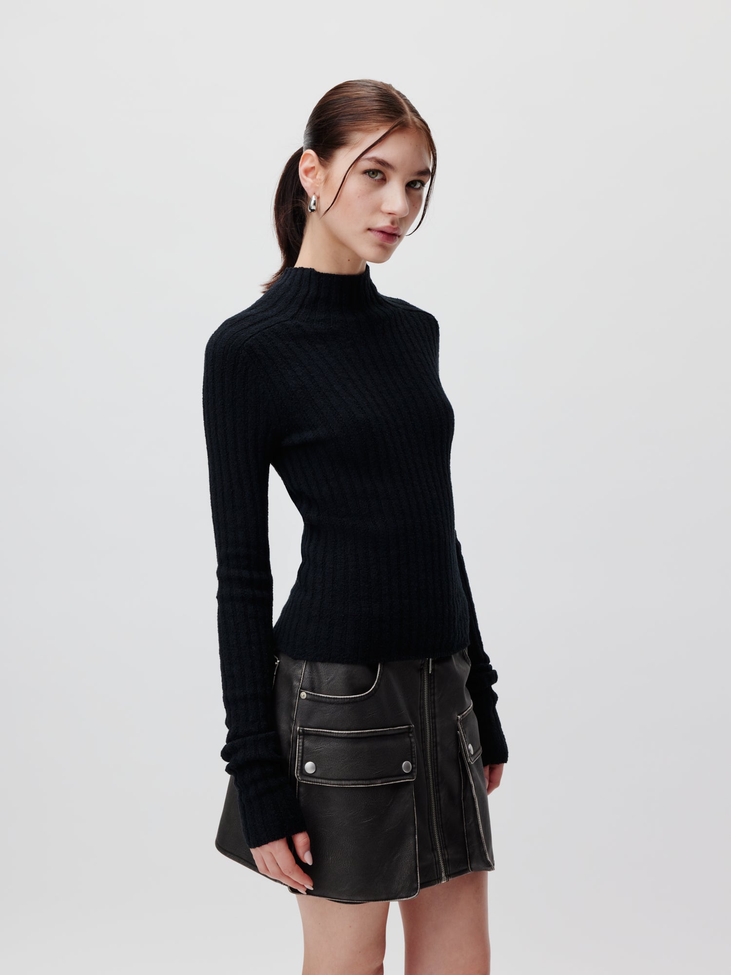 Eine Frau steht vor einem hellgrauen Hintergrund. Sie hat ihr Haar zurückgebunden und trägt einen taillierten, langärmeligen, schwarzen Rippenpullover namens „Franca“ von LeGer by Lena Gercke sowie einen schwarzen Lederrock mit Vordertaschen und Reißverschlussdetails. Die schmale Silhouette und die normale Länge des Ensembles verleihen ihm einen eleganten, modernen Look. Sie blickt leicht zur Seite und hat einen neutralen Gesichtsausdruck.