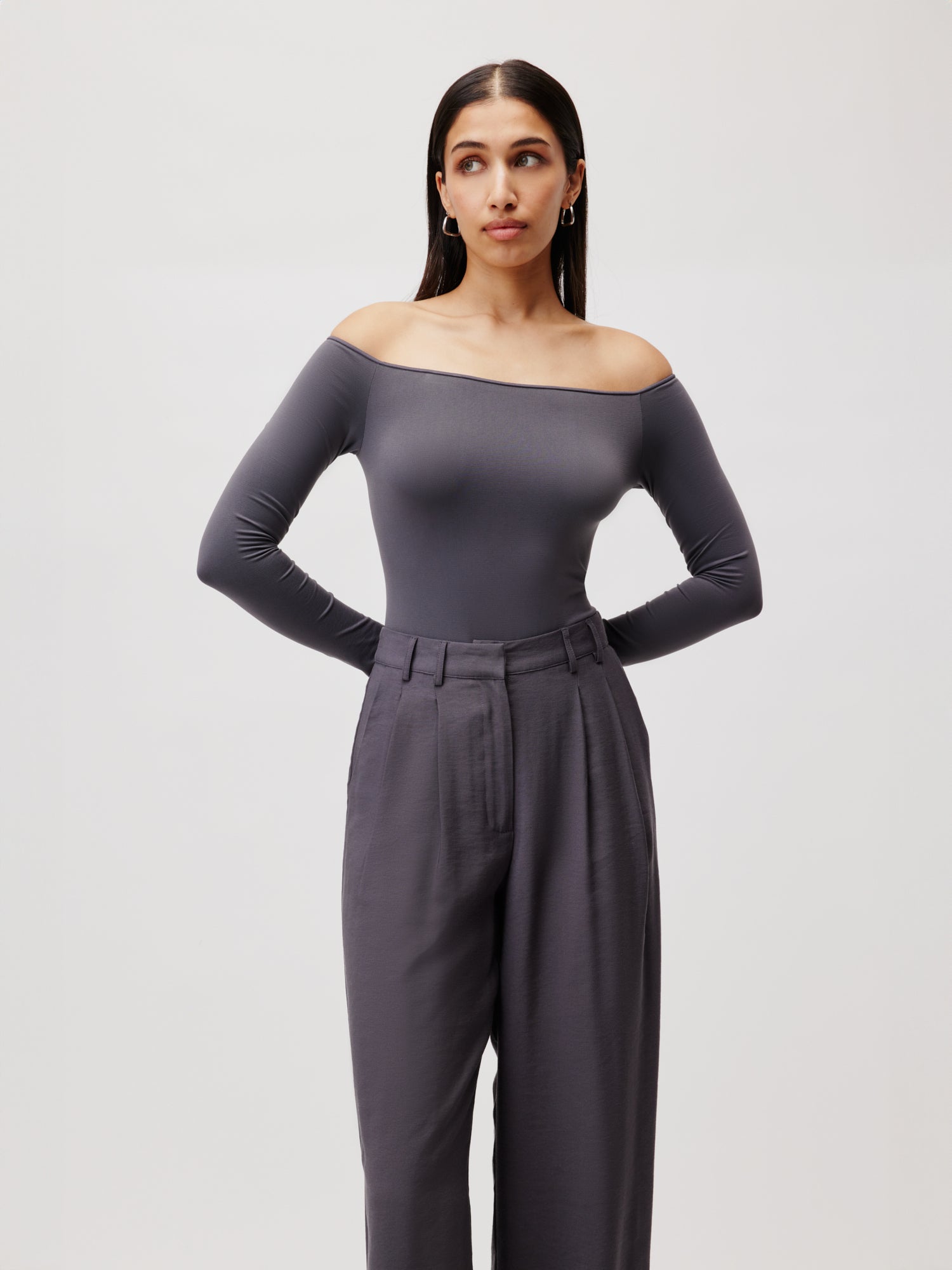 Eine Frau posiert vor einem schlichten Hintergrund. Sie trägt den grauen schulterfreien Body Athina von LeGer by Lena Gercke und eine graue Hose mit hoher Taille und stützt ihre Hände auf die Hüften.