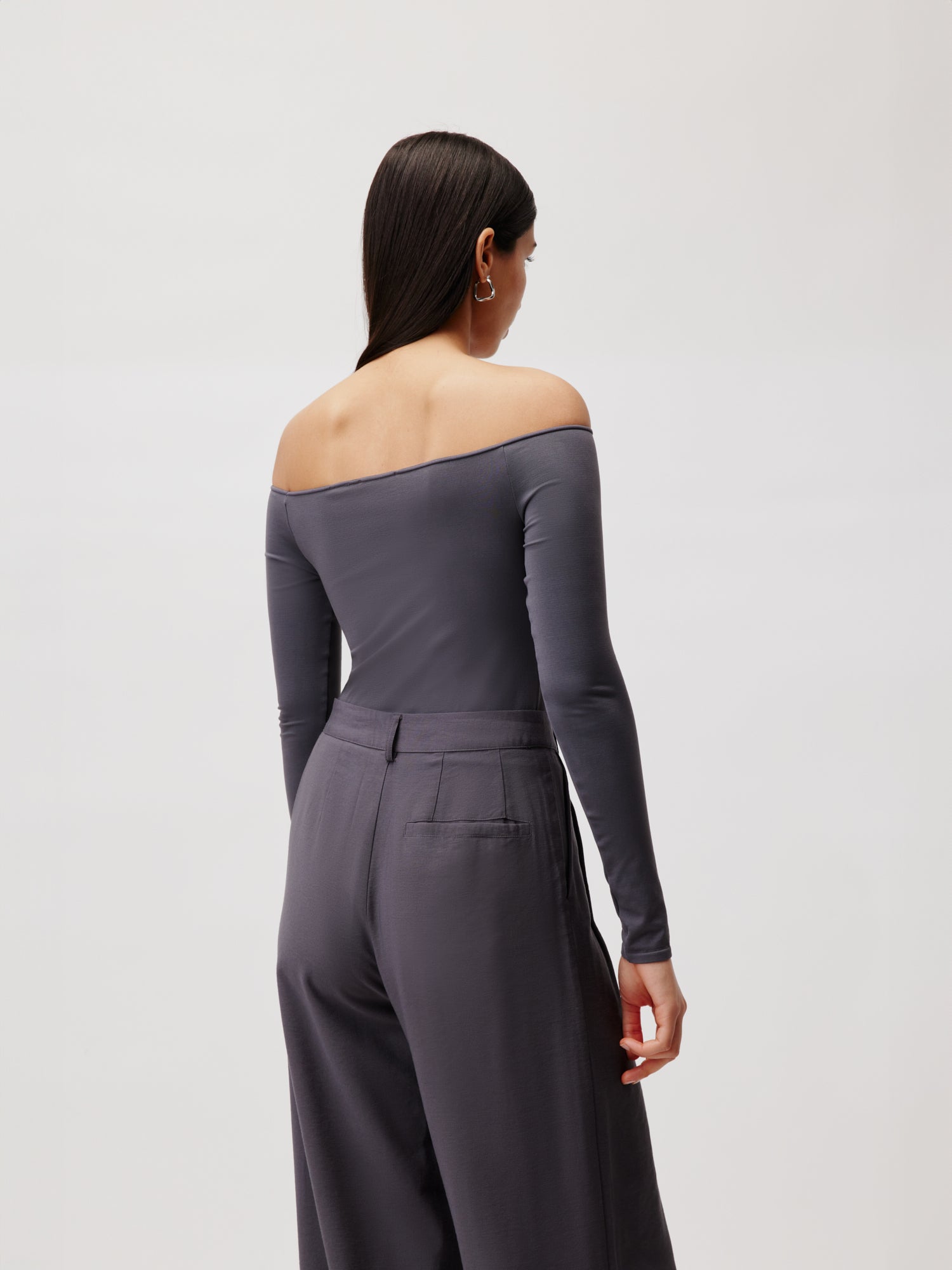 Eine Frau mit langen, dunklen Haaren blickt abgewandt in den „Body Athina“ von LeGer by Lena Gercke, einem schulterfreien Design in Dunkelgrau mit dazu passender Hose mit weitem Bein in normaler Länge. Vor einem hellgrauen Hintergrund wird ihr Look elegant eingerahmt, während der Langarm-Stil ihrer selbstbewussten Präsenz einen modernen Touch verleiht.