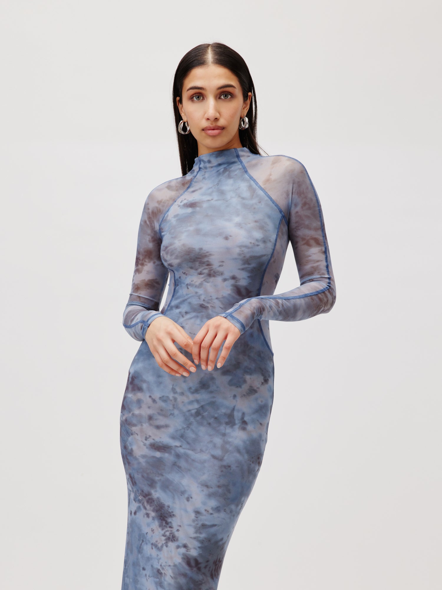 Eine Frau posiert vor einem hellgrauen Hintergrund und trägt das „Kleid Daliah“ von LeGer by Lena Gercke, ein schmal geschnittenes, 3/4-langes blaues Batikkleid mit Langarm und transparentem Stoff. Mit langen dunklen Haaren und Creolen blickt sie direkt in die Kamera.