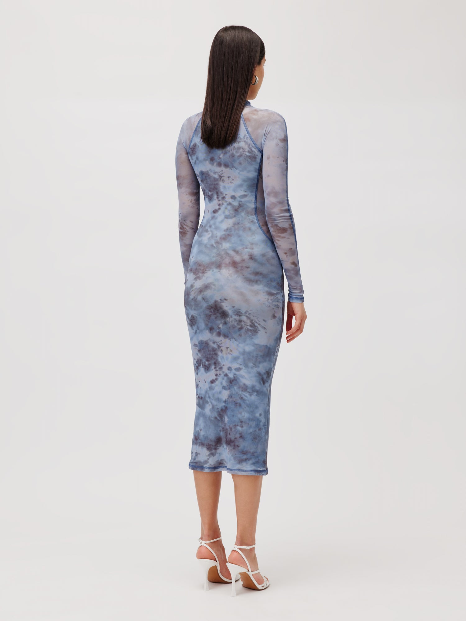 Eine Person steht mit dem Rücken zur Kamera und trägt ein schmales 3/4-langes Kleid Daliah von LeGer by Lena Gercke in blauem Batikmuster und weiße Riemchensandalen. Der Hintergrund ist schlicht und hellgrau.