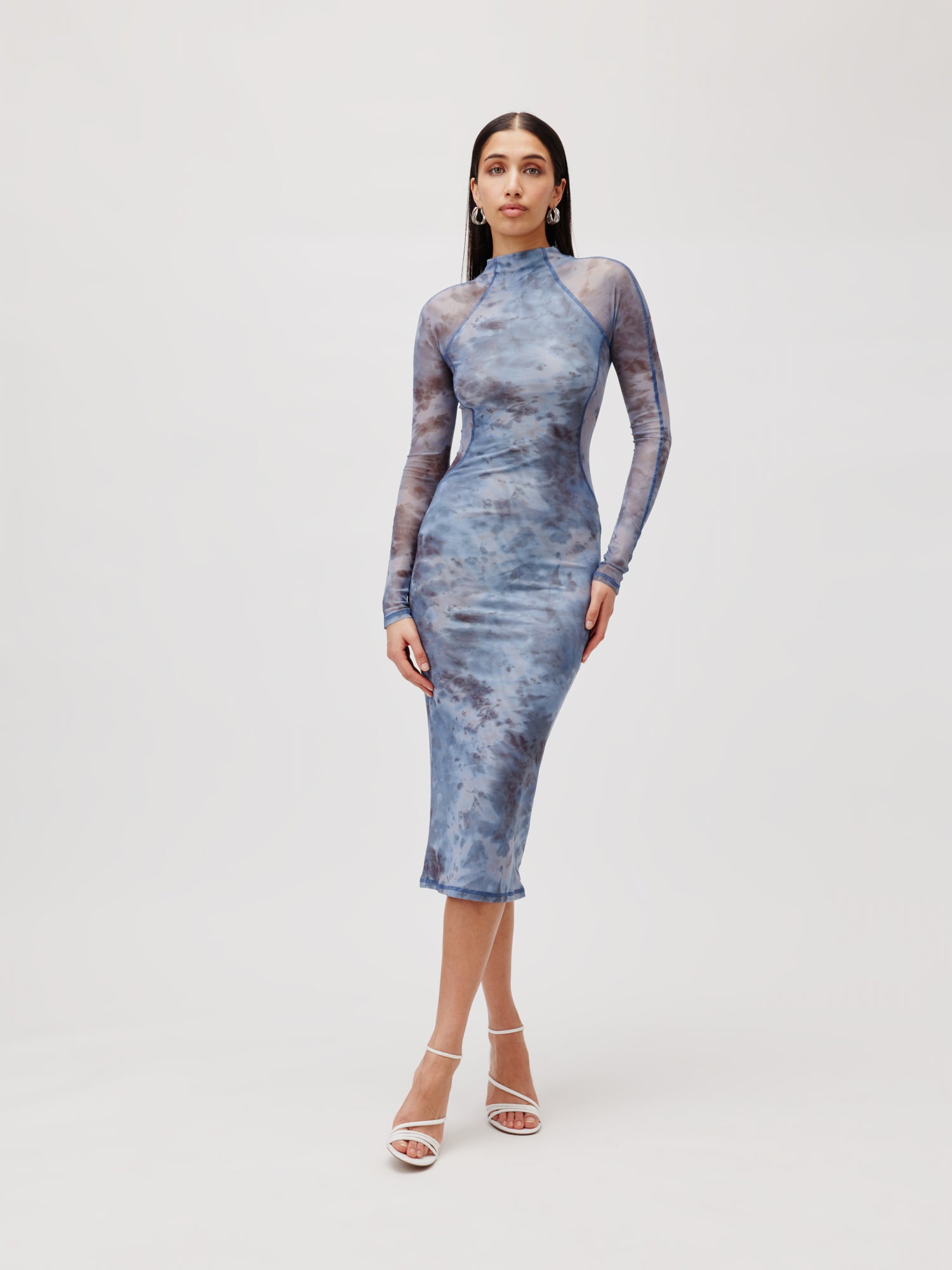 Eine Frau steht vor einem schlichten Hintergrund und trägt das figurbetonte Kleid Daliah, ein langärmliges blaues Batikkleid mit hohem Ausschnitt von LeGer by Lena Gercke. Sie vervollständigt das elegante Outfit mit weißen Riemchen-Pumps und großen Creolen. Ihr langes, dunkles Haar ist glatt gestylt.