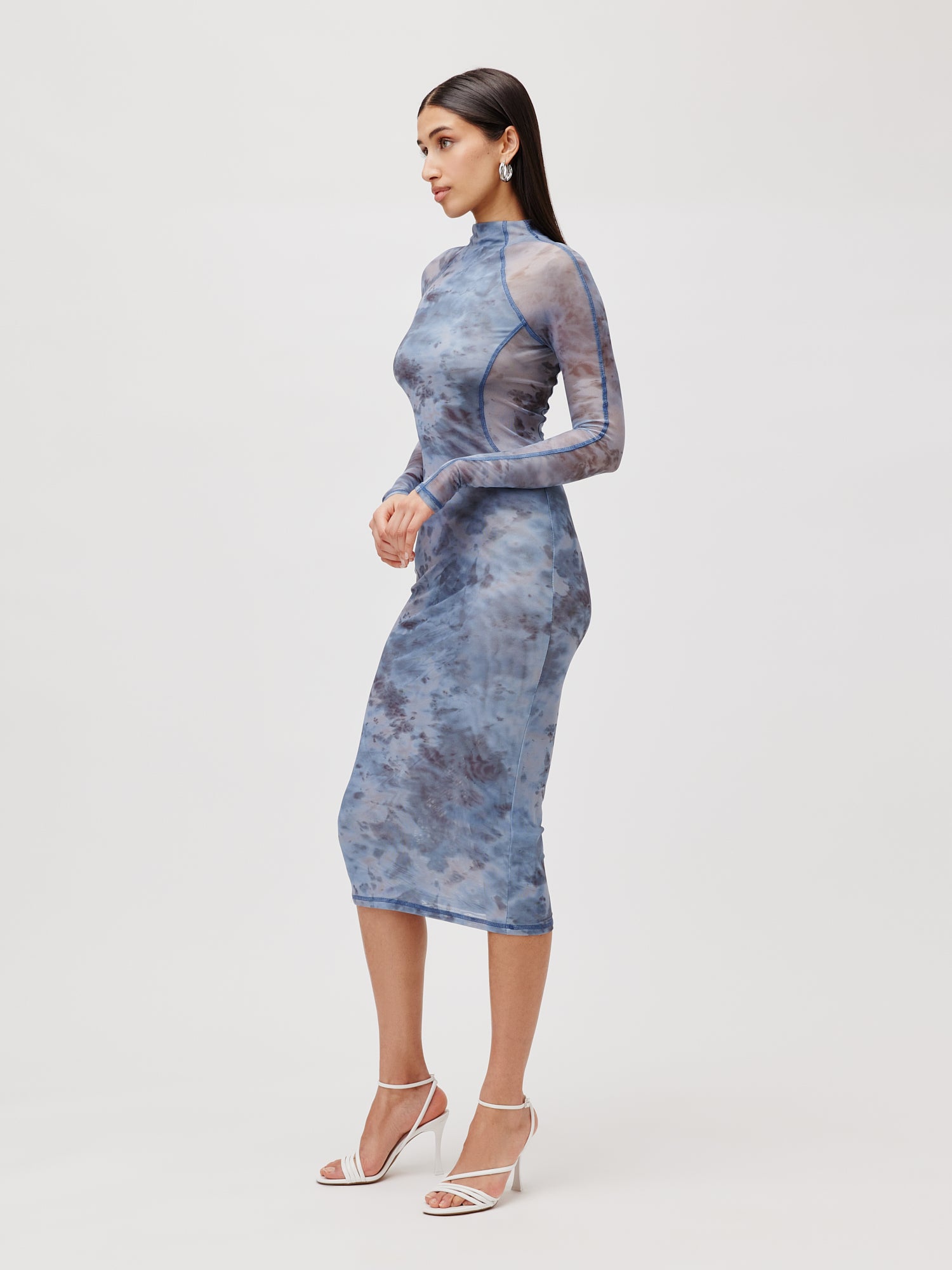 Eine Frau posiert elegant im halbtransparenten, 3/4-langen Kleid Daliah von LeGer by Lena Gercke mit einem marmorierten blauen Muster. Sie ergänzt das Kleid mit weißen High Heels und steht anmutig vor einem schlichten hellgrauen Hintergrund.