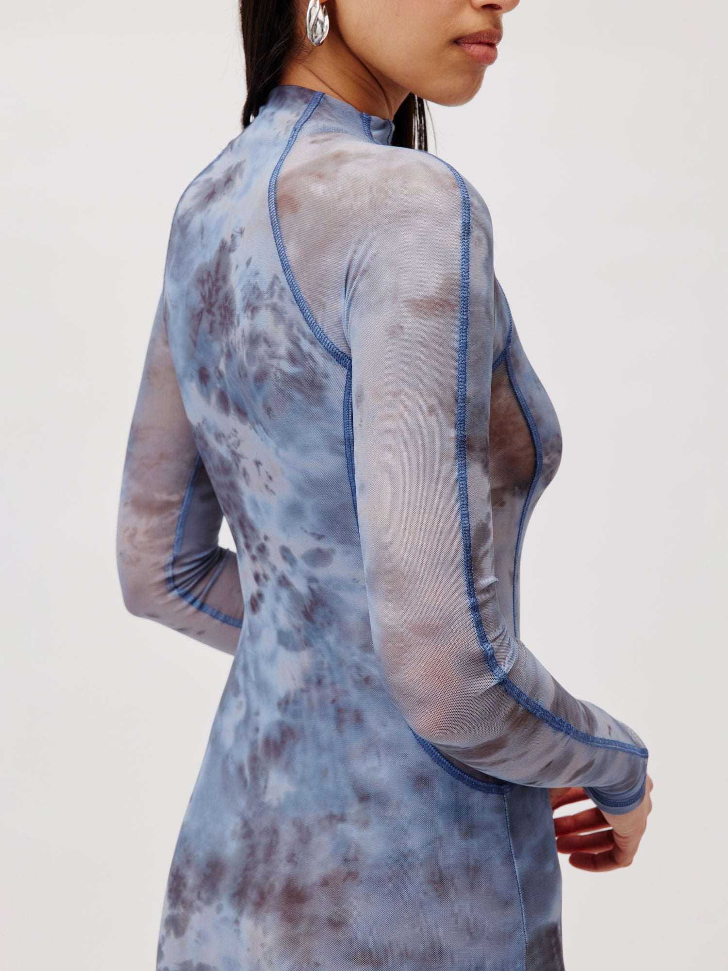 Eine Person trägt das Kleid Daliah von LeGer by Lena Gercke, ein langärmliges transparentes Kleid in Batiktönen in Blau und Grau. Das Langarmkleid hat einen hohen Halsausschnitt und sichtbare Nähte. Sie steht mit leicht angewinkeltem rechten Arm da und zeigt einen Teil ihres Gesichts und Haares.