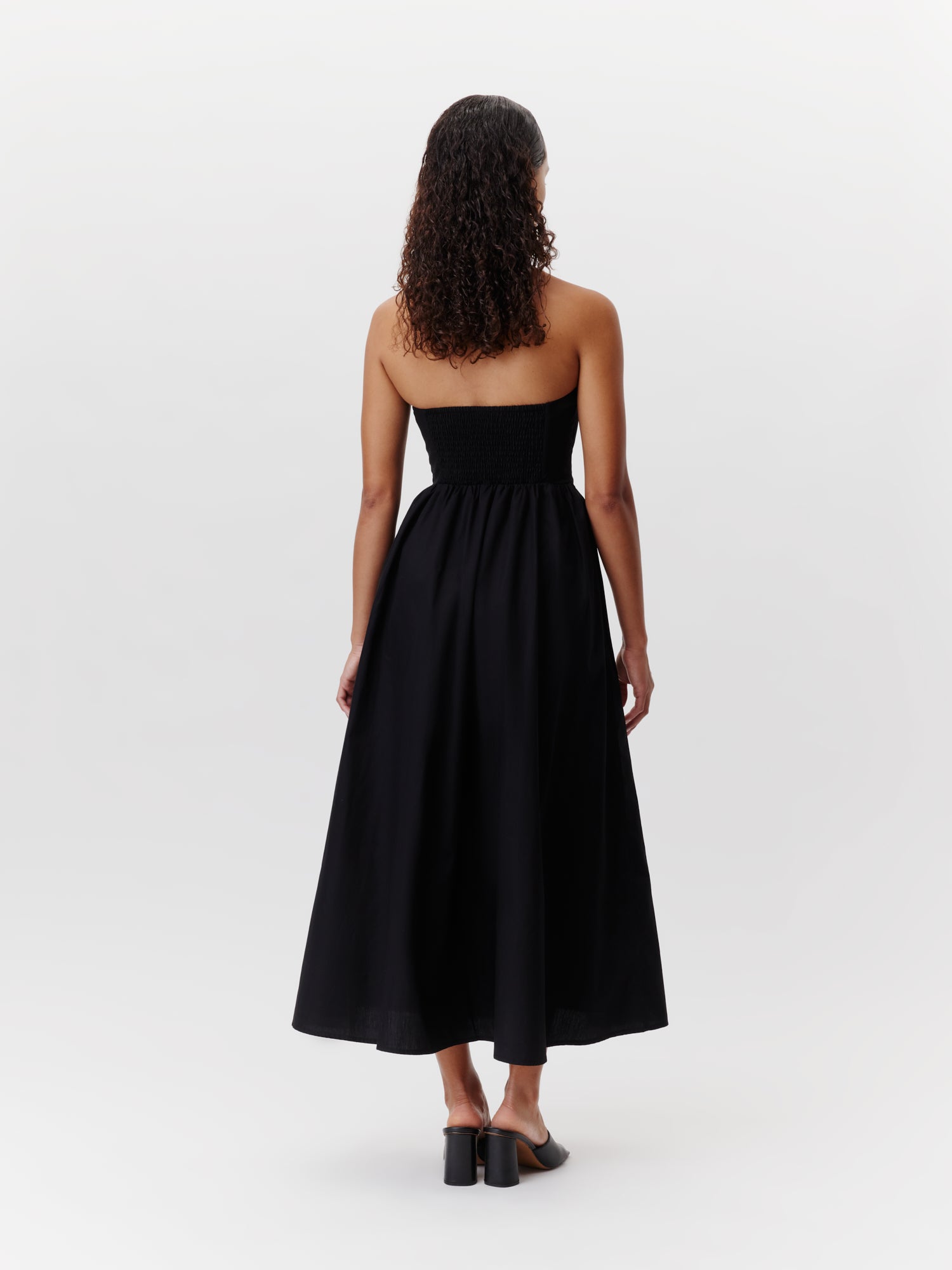 Eine Frau mit lockigem Haar trägt das Vianne-Kleid von LeGer by Lena Gercke, ein trägerloses, bodenlanges schwarzes Kleid in Größe 36, kombiniert mit schwarzen Schuhen mit Absatz. Sie blickt abgewandt vor einem schlichten weißen Hintergrund.