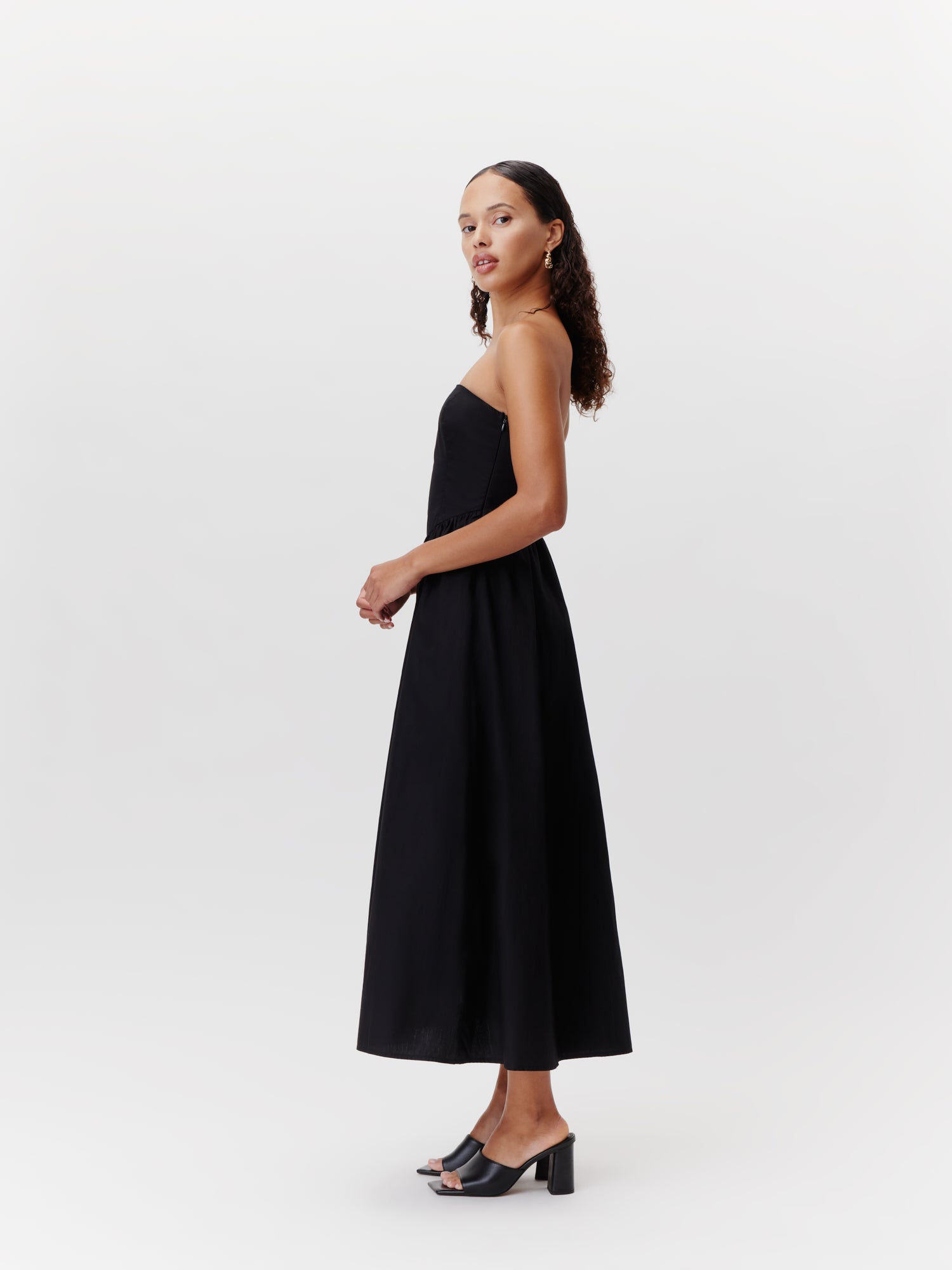 Eine Frau mit lockigem Haar steht seitlich im Kleid „Vianne“ von LeGer by Lena Gercke, einem schwarzen, trägerlosen Modell in 7/8-Länge, kombiniert mit schwarzen Sandalen mit Absatz, vor einem schlichten weißen Hintergrund.