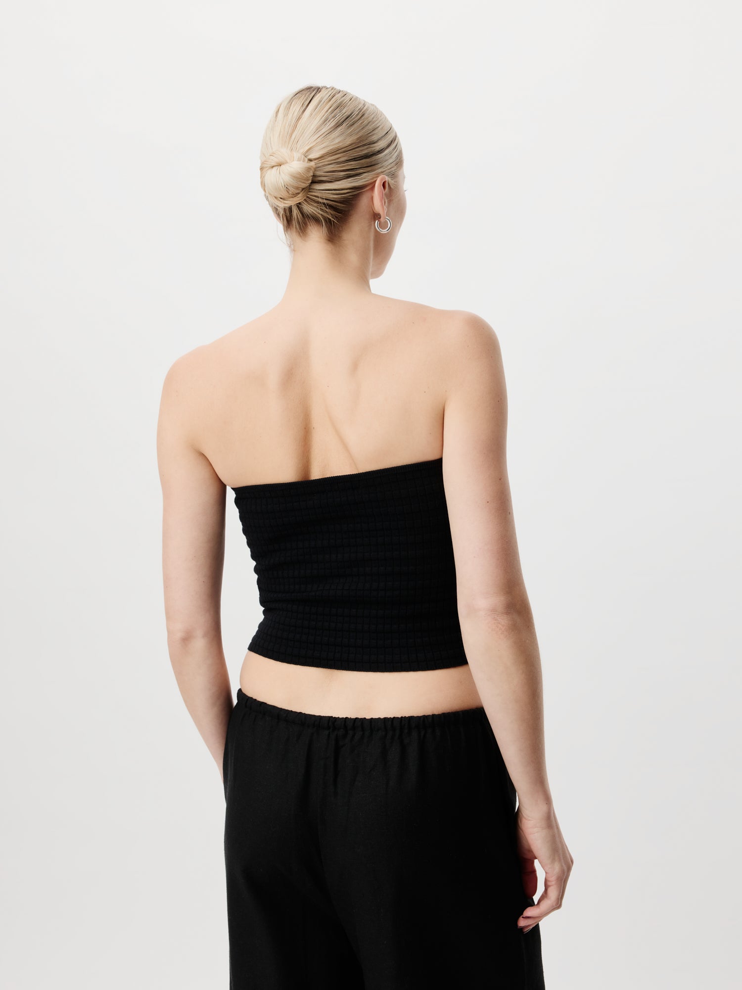 Eine Person mit blondem Haar zu einem Dutt, die ein schmales, ärmelloses schwarzes Cathleen-Top von LeGer by Lena Gercke und eine kurze schwarze Hose trägt, steht mit dem Rücken zur Kamera vor einem schlichten weißen Hintergrund.