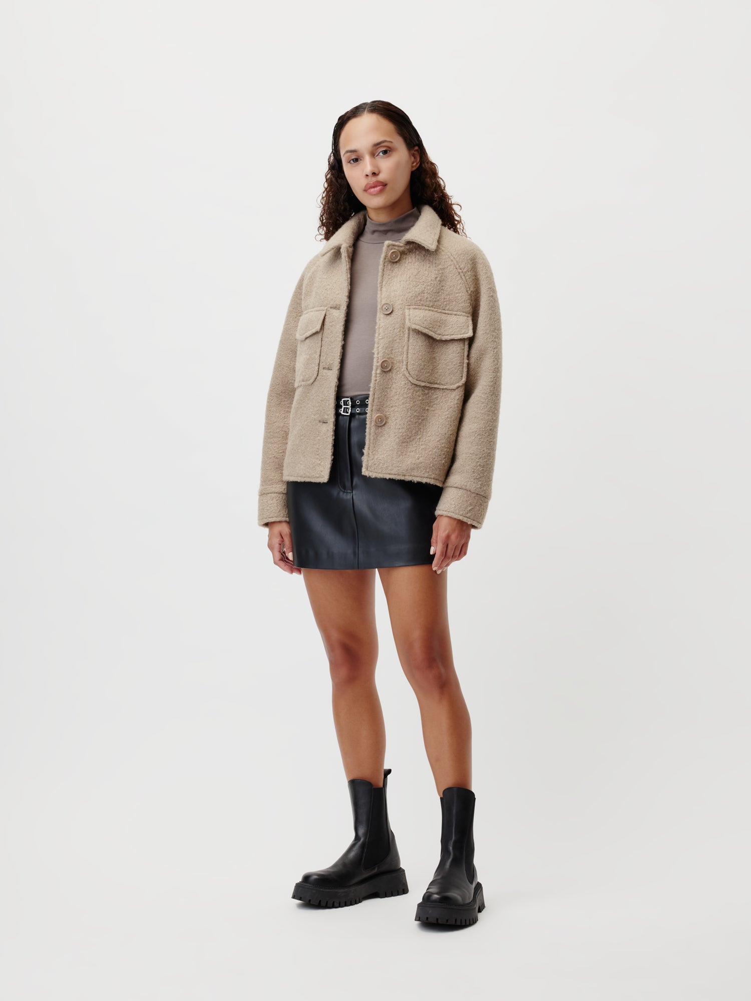 Eine Frau mit lockigem Haar und neutralem Gesichtsausdruck steht vor einem weißen Hintergrund und trägt die beigefarbene Maura Jacket von LeGer by Lena Gercke über einem taupefarbenen Oberteil, einem schwarzen Lederminirock und schwarzen Ankle Boots.
