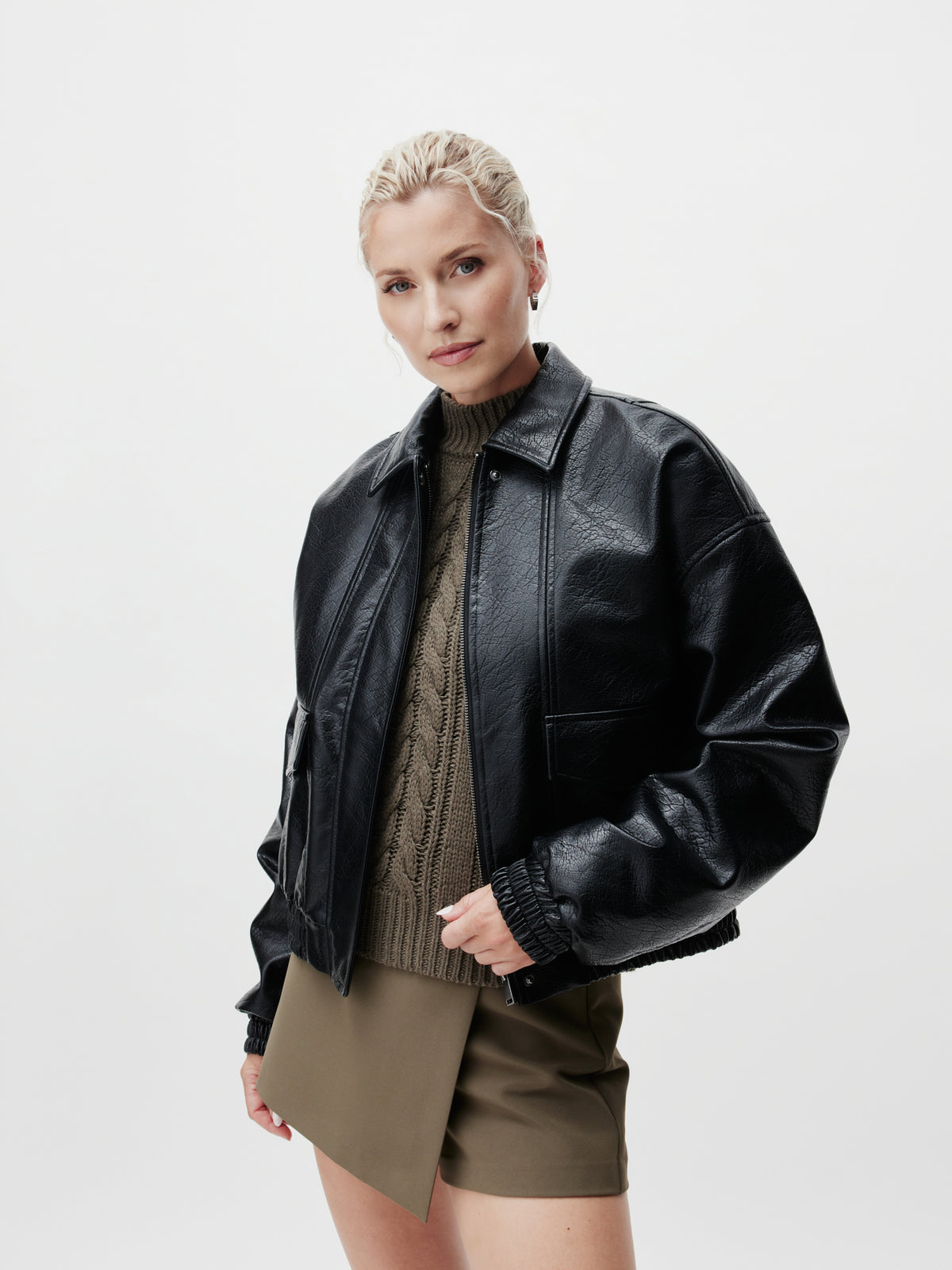 Eine Frau mit blonden, zu einem Dutt zusammengebundenen Haaren trägt die schwarze „Taira“-Langarm-Lederjacke von LeGer by Lena Gercke in normaler Länge über einem braunen Zopfmusterpullover und einem olivgrünen Rock in Größe 36, während sie vor einem schlichten weißen Hintergrund steht.