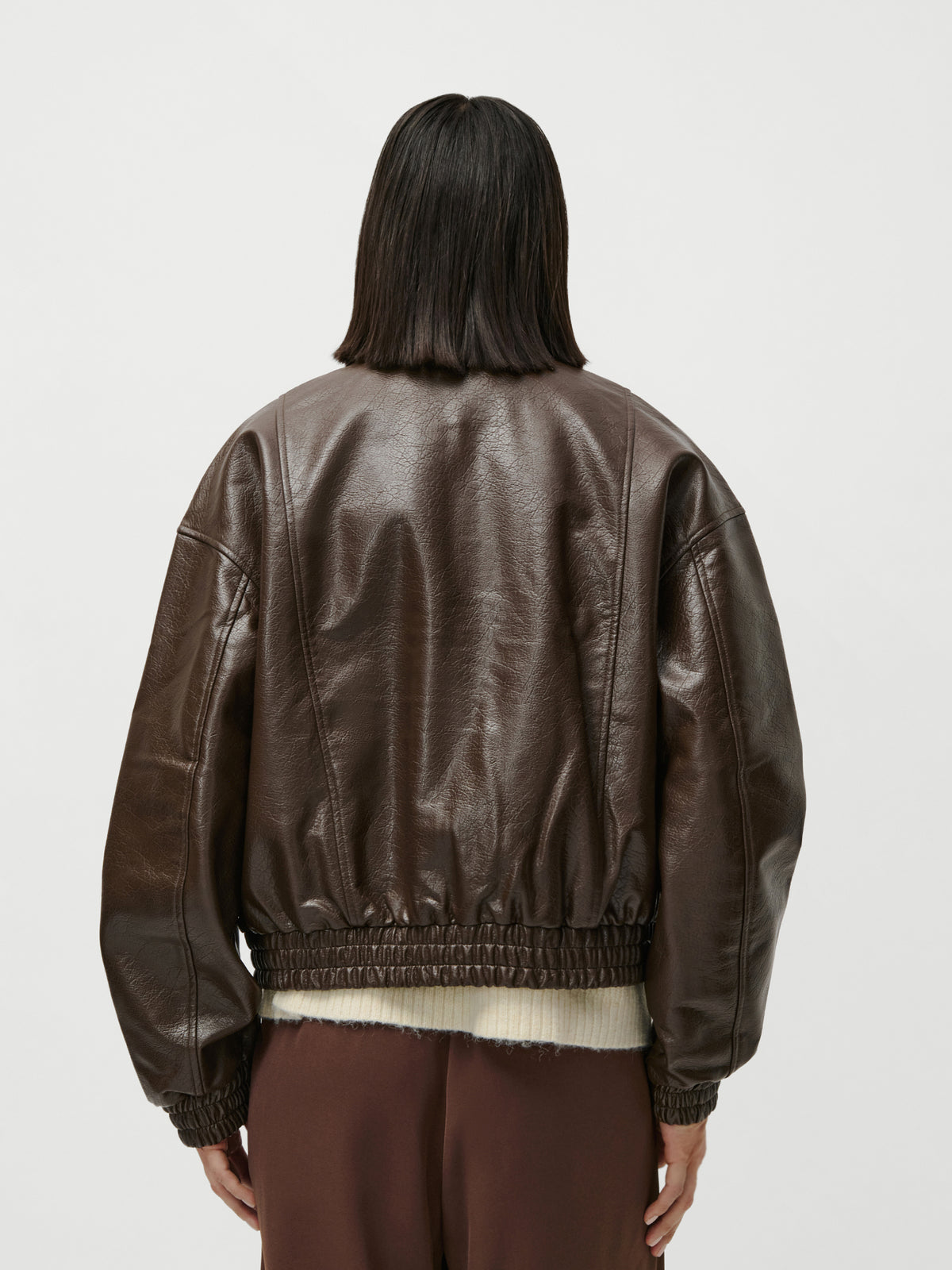 Eine Person mit glattem, schulterlangem, dunklem Haar wird von hinten vor einem schlichten weißen Hintergrund fotografiert. Sie trägt die Jacke „Taira“ von LeGer by Lena Gercke – eine locker sitzende, dunkelbraune Lederjacke mit langen Ärmeln –, dazu eine braune Hose in Größe 36 und normaler Länge.