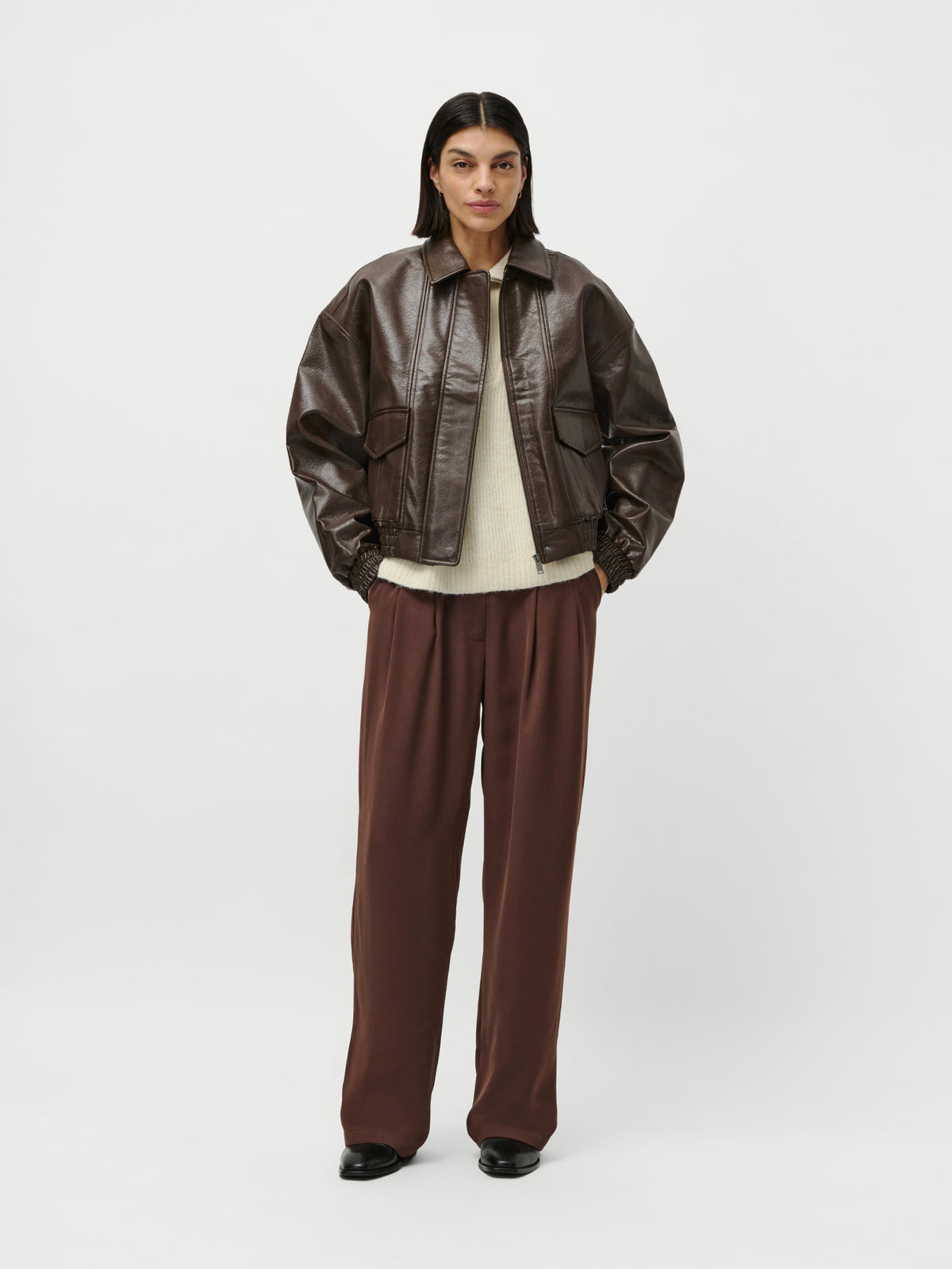 Eine Person steht vor einem schlichten Hintergrund und trägt die braune Lederjacke „Taira“ von LeGer by Lena Gercke in normaler Länge, einen cremefarbenen Langarmpullover, eine lockere braune Hose in Größe 36 und schwarze Schuhe; die Hände stecken in den Taschen, der Gesichtsausdruck ist neutral.