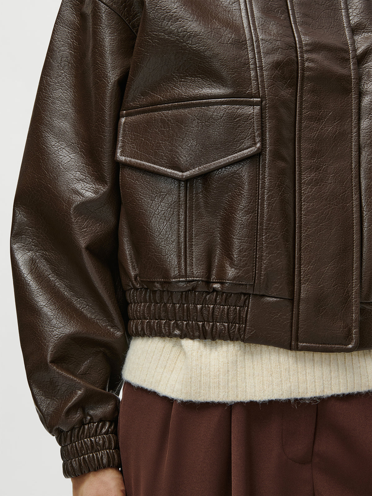 Eine Nahaufnahme einer Person, die die dunkelbraune Lederjacke „Taira“ von LeGer by Lena Gercke mit Patten-Taschen trägt, kombiniert mit einem cremefarbenen, gerippten Langarmpullover und einer braunen Hose in Größe 36, alles in normaler Länge.