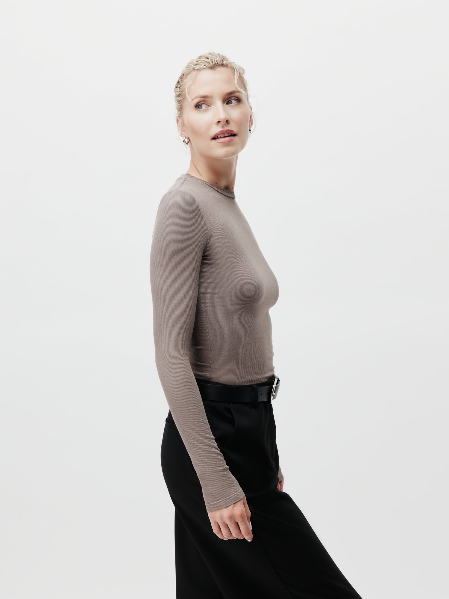 Eine Frau mit blondem Haar in einem tiefen Zopf trägt das taillierte Cinzia Longsleeve von LeGer by Lena Gercke in Taupe, gepaart mit einer schwarzen Hose. Sie steht seitlich vor einem weißen Hintergrund. Kaufen Sie dieses Modell im LeGer Onlineshop.