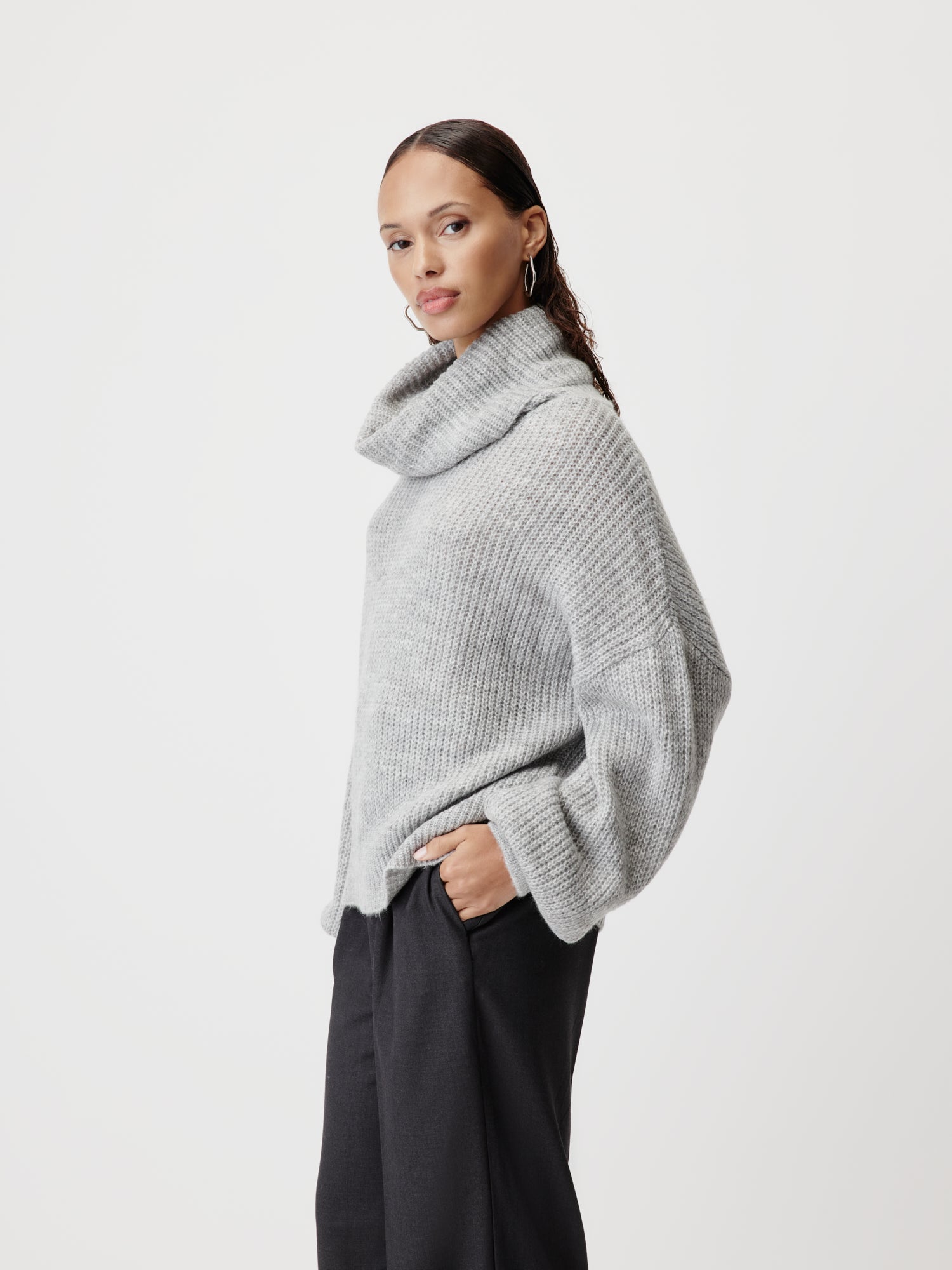 Eine Frau steht vor einem schlichten Hintergrund und trägt den Jarine Jumper von LeGer by Lena Gercke - einen lockeren, hellgrauen Rollkragenpullover mit extralangen Ärmeln und normaler Länge - zu einer dunklen Hose mit weitem Bein, eine Hand in der Tasche.