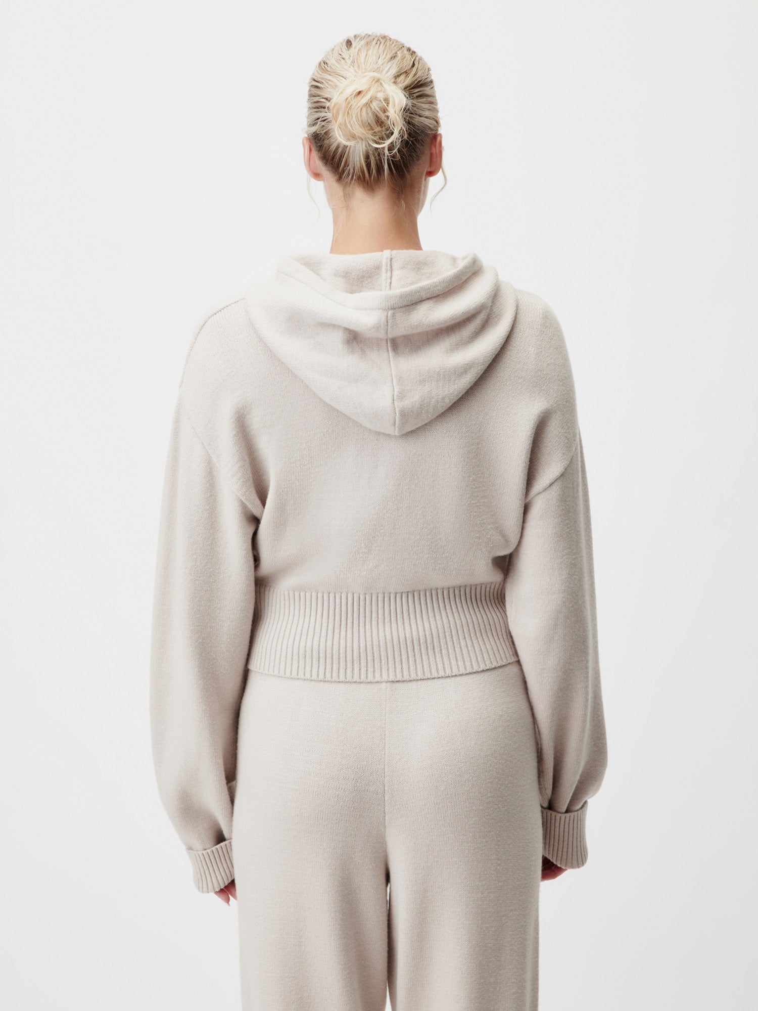 Eine Frau mit blonden Haaren in einem Dutt ist von hinten zu sehen. Sie trägt den LeGer by Lena Gercke Cornelia Cardigan in Größe 36, langarm hellbeige, gepaart mit einer passenden Hose. Das weiche, gemütliche Outfit hebt sich von einem schlichten weißen Hintergrund ab.