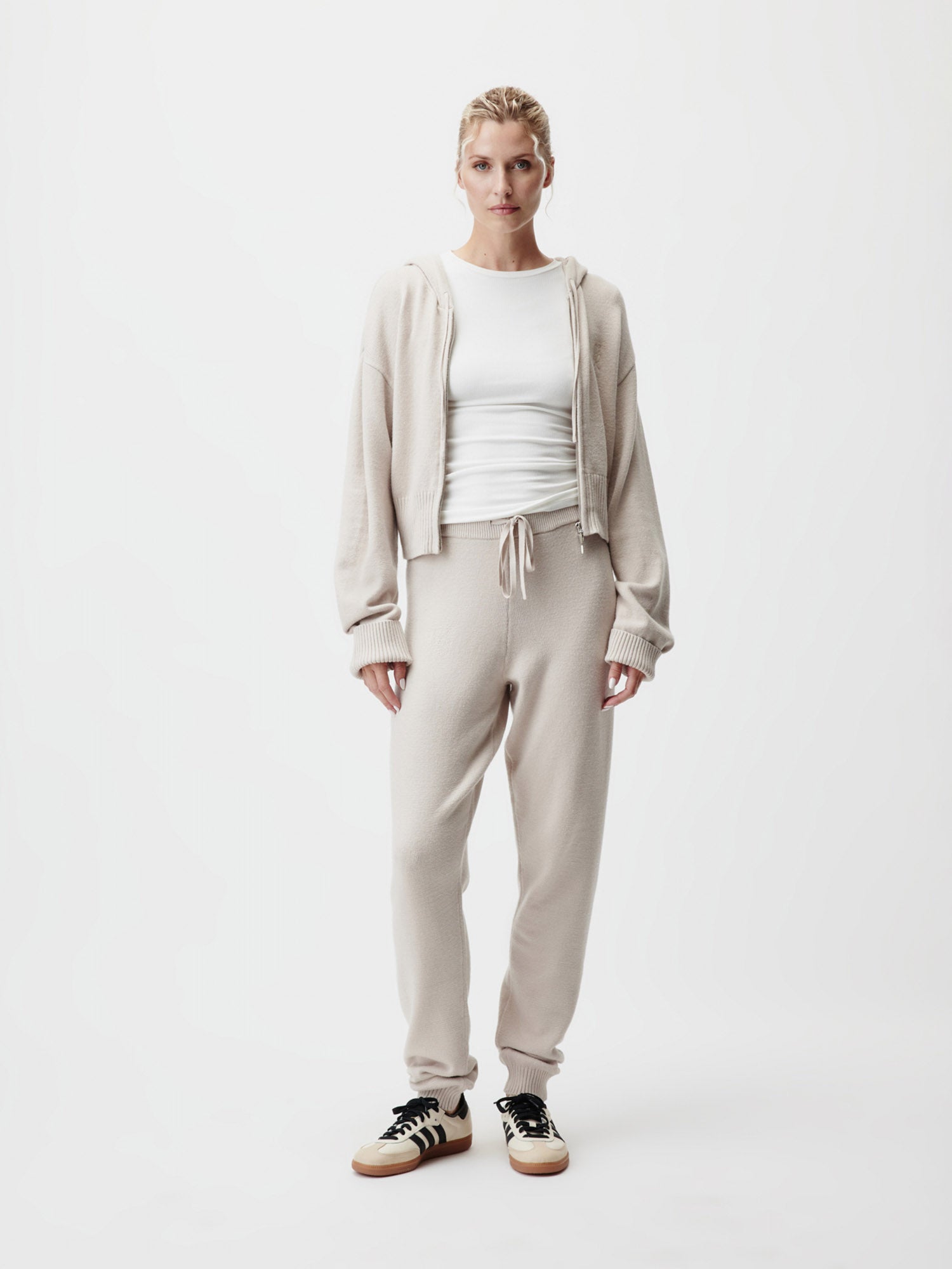 Eine Frau blickt nach vorne und trägt eine cremefarbene Cornelia-Strickjacke von LeGer by Lena Gercke in Größe 36, eine passende Jogginghose mit Tunnelzug, ein weißes Hemd und schwarz-weiße Turnschuhe vor einem schlichten weißen Hintergrund.