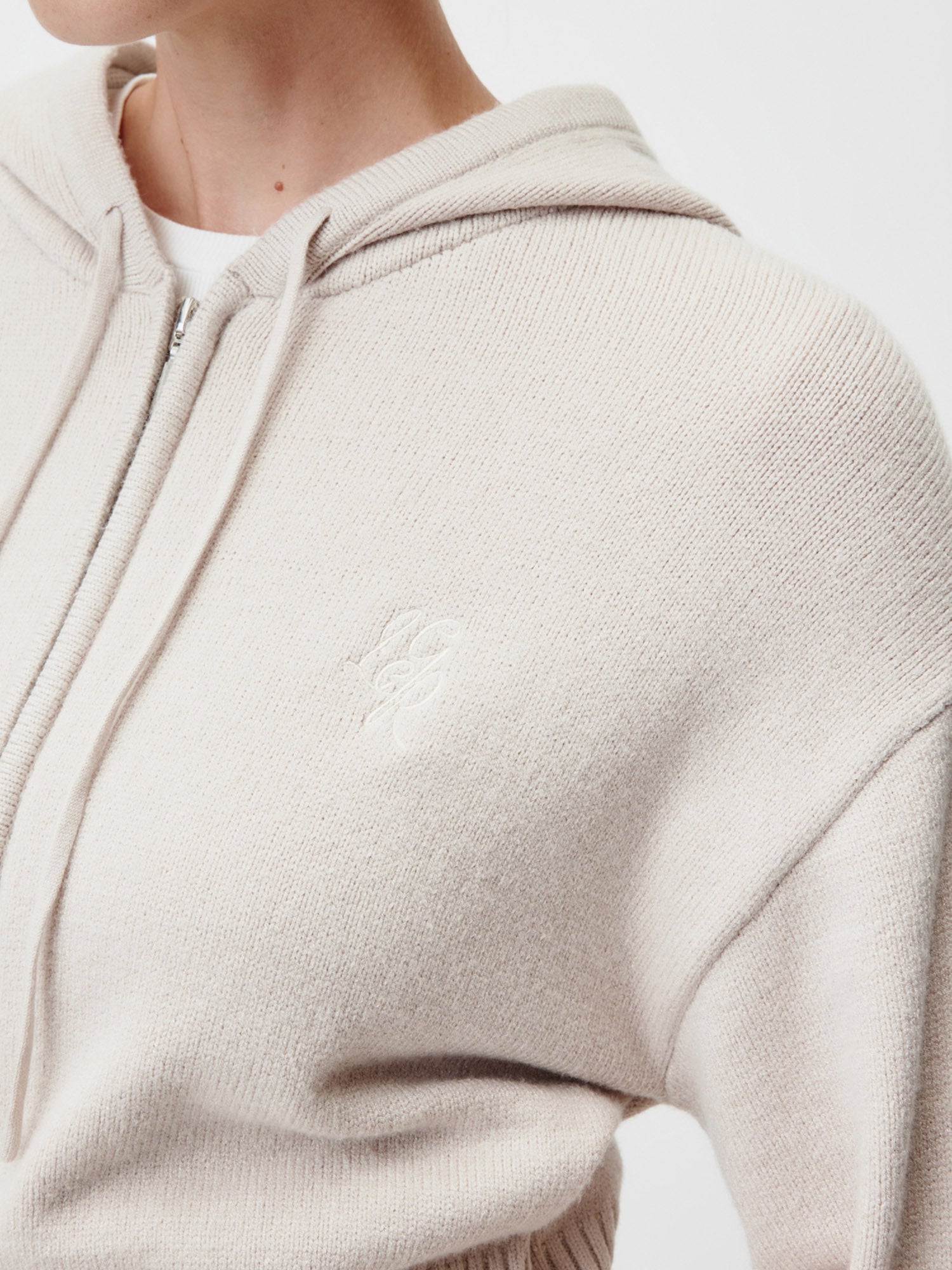 Nahaufnahme einer Person, die den LeGer by Lena Gercke Cornelia Cardigan in Hellbeige, Größe 36, mit einem dezenten Brustlogo trägt. Die Strickjacke hat einen Frontreißverschluss, Kordelzüge und eine normale Länge. Nur ein Teil des Gesichts und des Oberkörpers ist sichtbar.