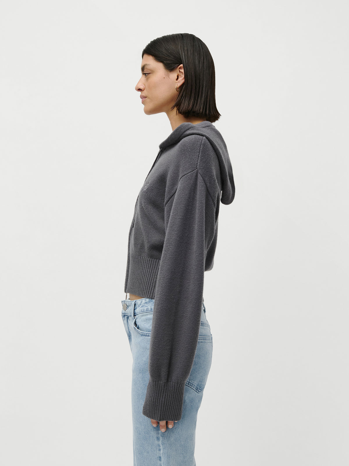 Eine Person mit glattem, dunklem Haar im Bob-Schnitt steht im Profil vor einem schlichten weißen Hintergrund und trägt den Cardigan „Cornelia“ von LeGer by Lena Gercke – einen grauen Loungewear-Pullover mit Kapuze –, kombiniert mit hellblauen Jeans.