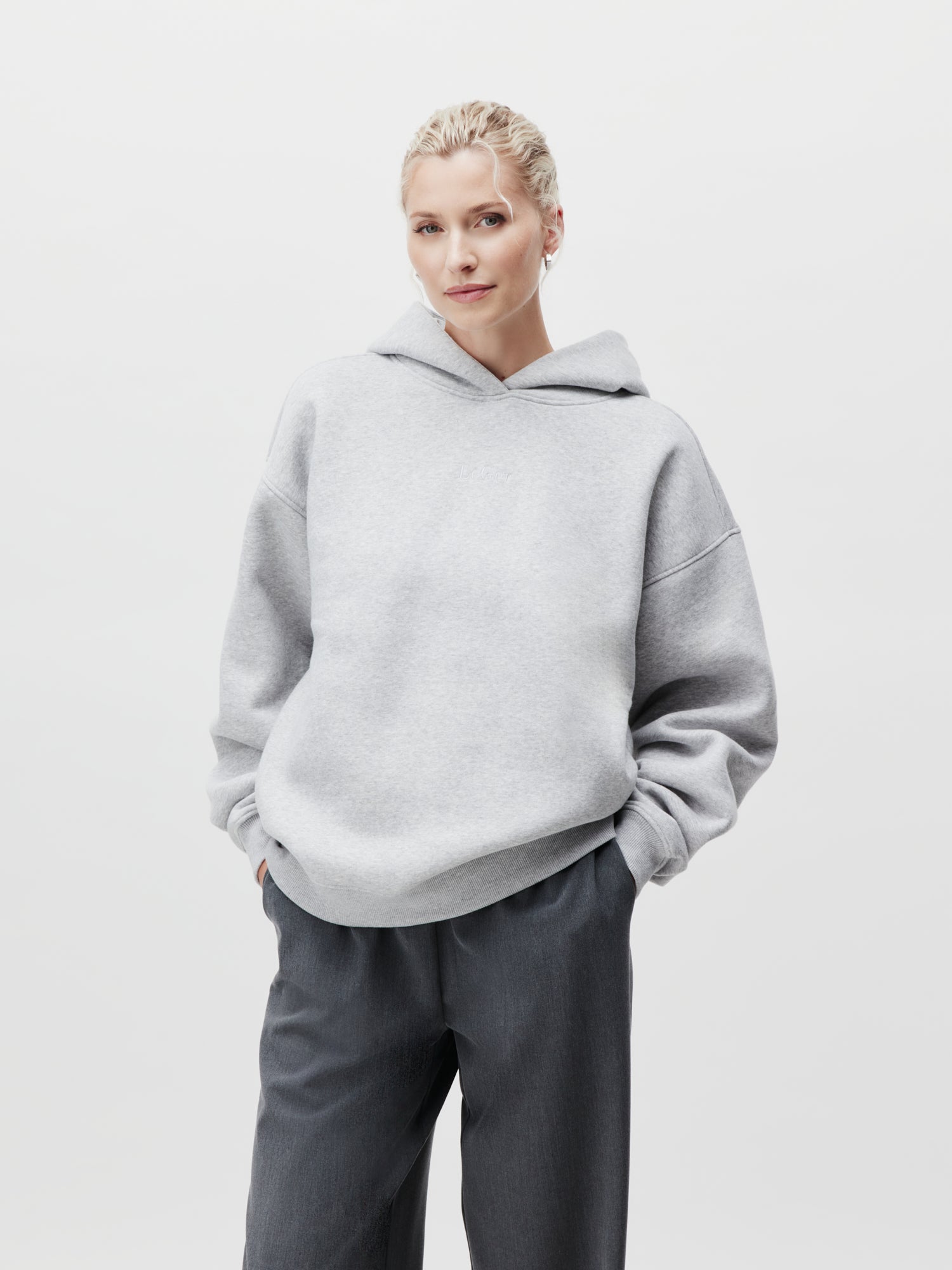 Eine Person steht vor einem schlichten Hintergrund und trägt einen hellgrauen Abby Hoodie mit langen Ärmeln von LeGer by Lena Gercke und eine dunkelgraue Hose. Sie hat kurze, helle Haare und ihre Hände sind in die Kapuzentaschen gesteckt. Das Outfit hat Größe 36 und bietet eine bequeme Passform für den Alltag.