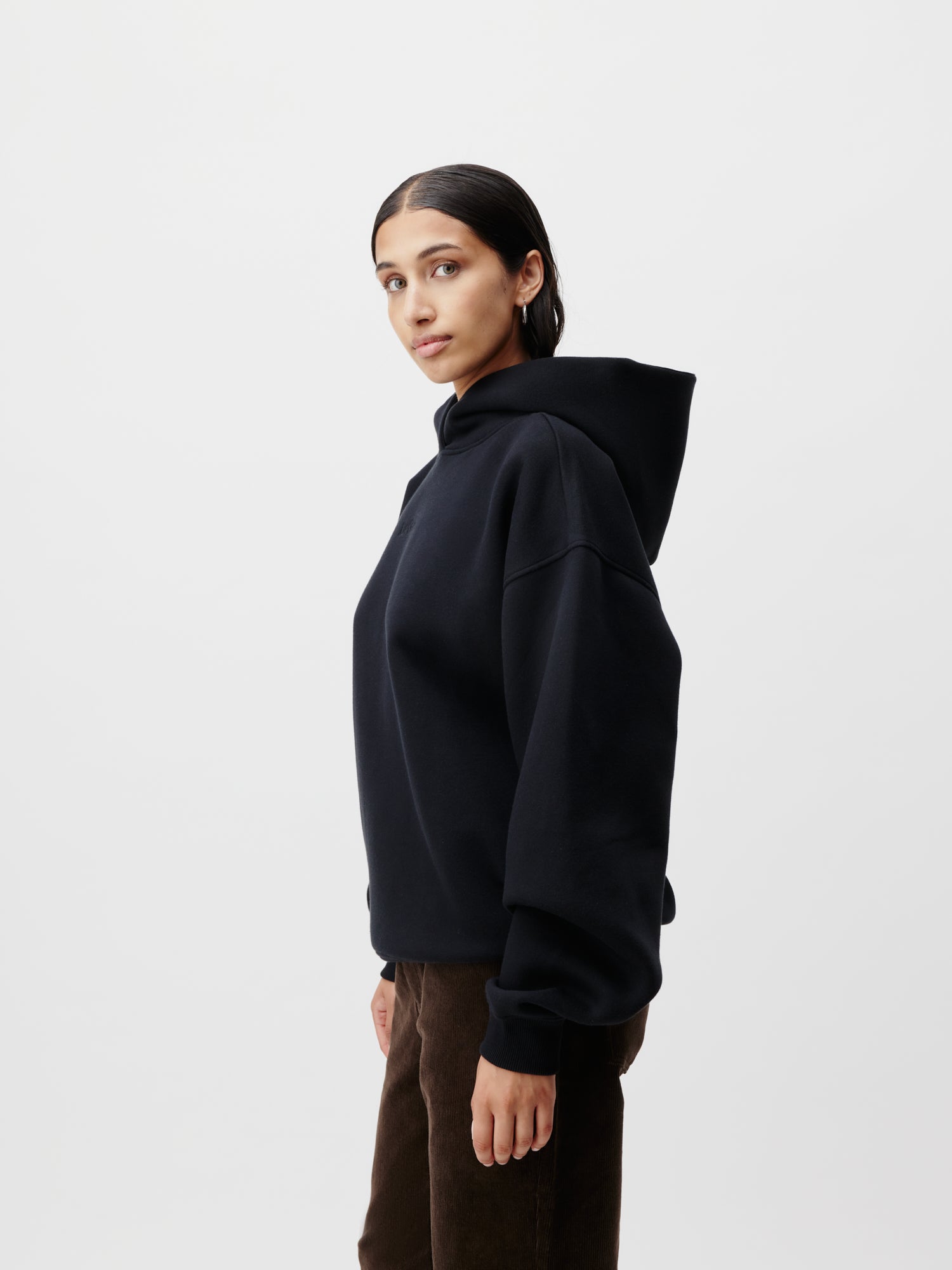 Eine Person mit langen dunklen Haaren steht seitlich, trägt einen schwarzen Abby Hoodie von LeGer by Lena Gercke und eine braune Hose in Größe 36 vor einem schlichten weißen Hintergrund. Sie blickt leicht über die Schulter.