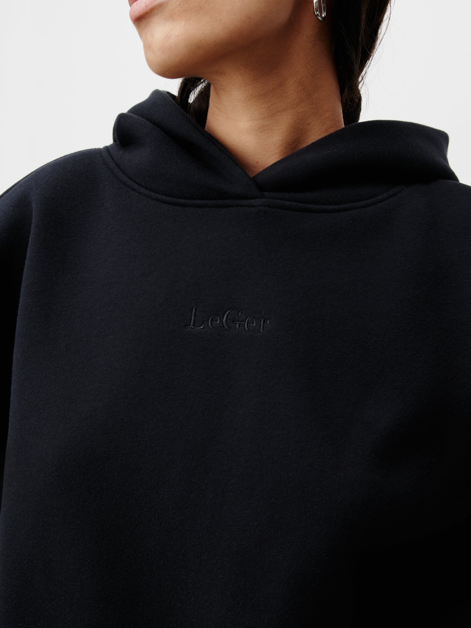 Eine Nahaufnahme einer Person, die einen schwarzen Abby Hoodie von LeGer by Lena Gercke trägt, auf dessen Vorderseite dezent „LeGer“ eingeprägt ist. Sie blickt zur Seite, wodurch die normale Passform der Größe 36 vor einem schlichten weißen Hintergrund hervorgehoben wird.