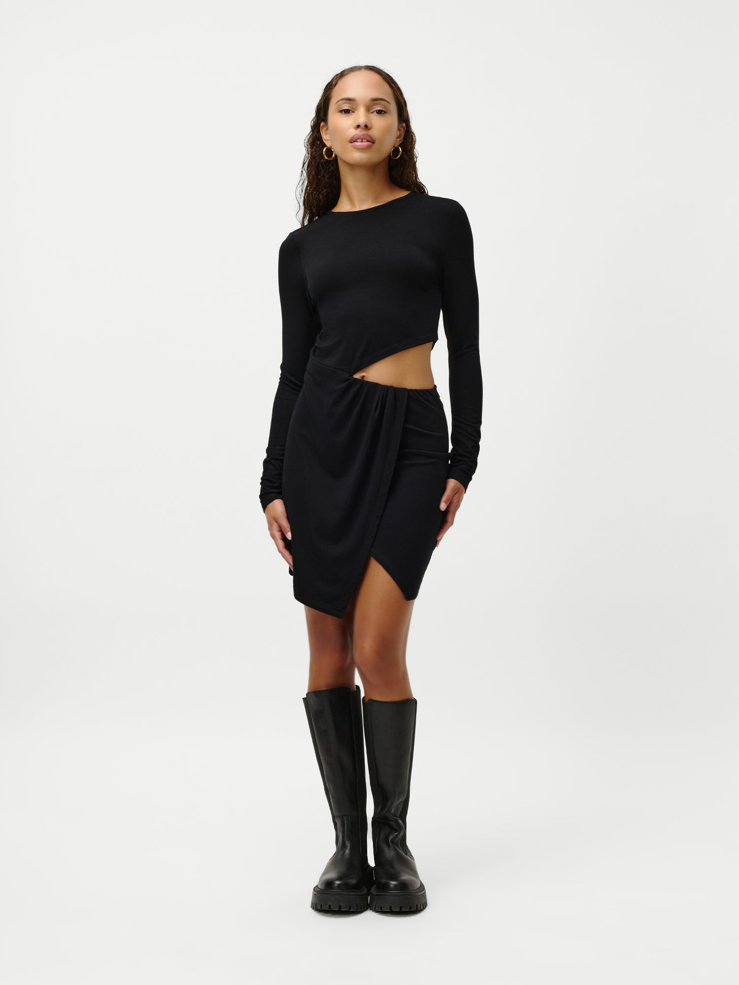 Eine Frau blickt nach vorne und trägt das Kleid „Farina“ von LeGer by Lena Gercke – ein schwarzes, langärmeliges Minikleid mit asymmetrischem Ausschnitt an der Taille und drapiertem Rock, kombiniert mit kniehohen schwarzen Stiefeln. Der Hintergrund ist schlicht weiß.