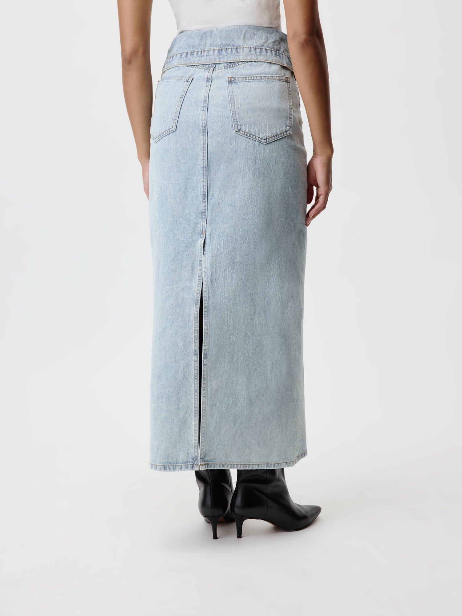 Sie tragen den Blanca-Rock von LeGer by Lena Gercke, ein hellblaues 7/8-Jeansteil mit Gesäßtaschen und Schlitz auf der Rückseite und normaler Passform. Dazu kombinieren sie nahtlos ein weißes Oberteil und schwarze Stiefel mit spitzer Spitze und stehen dabei mit dem Rücken zum Betrachter.