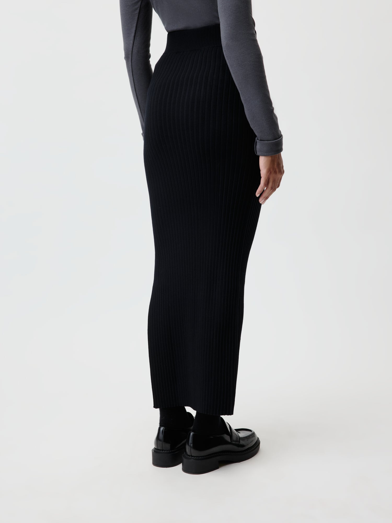 Eine Person trägt den Elenie-Rock von LeGer by Lena Gercke, einen langen schwarzen Rippenrock mit eleganter Passform, kombiniert mit einem dunklen Langarmshirt, schwarzen Socken und Schuhen. Das Ensemble hebt sich vom hellen Hintergrund ab.