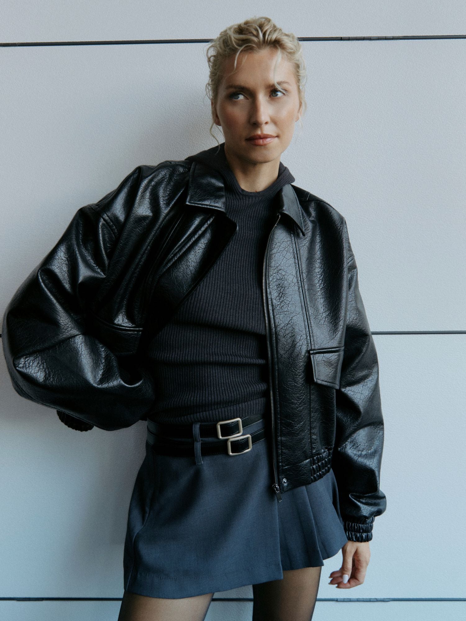 Eine blondhaarige Frau mit einer lockeren Hochsteckfrisur posiert vor einer weißen Wand. Sie trägt eine glänzende schwarze Lederjacke, ein geripptes schwarzes Oberteil, den Rock Lucca von LeGer by Lena Gercke mit Mini-Gürtel und normaler Passform sowie eine schwarze Strumpfhose.