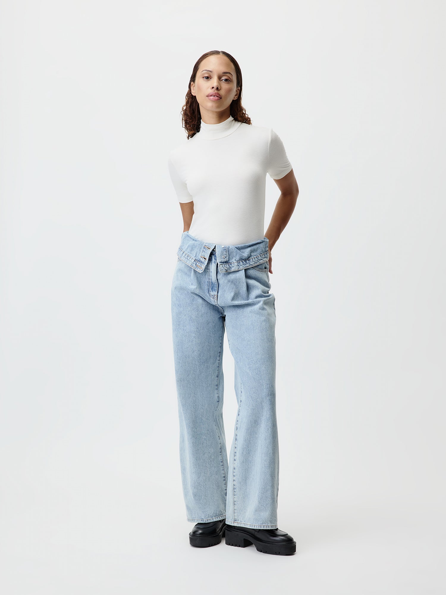 Eine Person steht vor einem schlichten Hintergrund und trägt ein weißes Rollkragenshirt und hoch taillierte, hellblaue Donia Pants von LeGer by Lena Gercke mit einem einzigartigen umgeschlagenen Hosenbund. Sie trägt außerdem schwarze Plateauschuhe, um etwas Länge hinzuzufügen. Ihre Hände sind hinter ihrem Rücken.