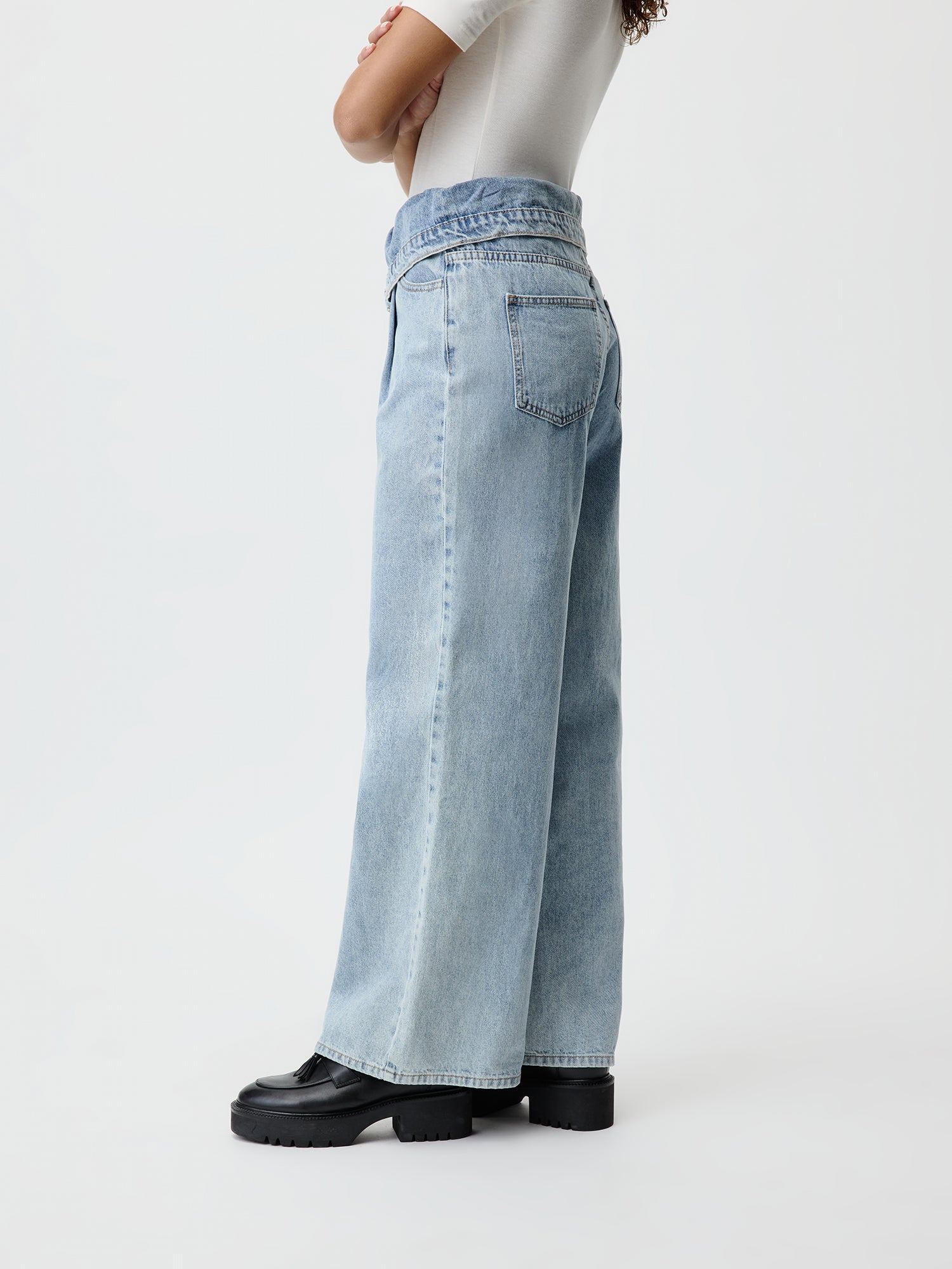 Eine Person trägt die Donia Pants von LeGer by Lena Gercke, eine hellblaue, hoch taillierte Jeans mit weitem Bein und Maxilänge, kombiniert mit einem weißen Hemd. Sie steht seitlich mit verschränkten Armen und trägt schwarze Stiefeletten vor einem schlichten weißen Hintergrund.