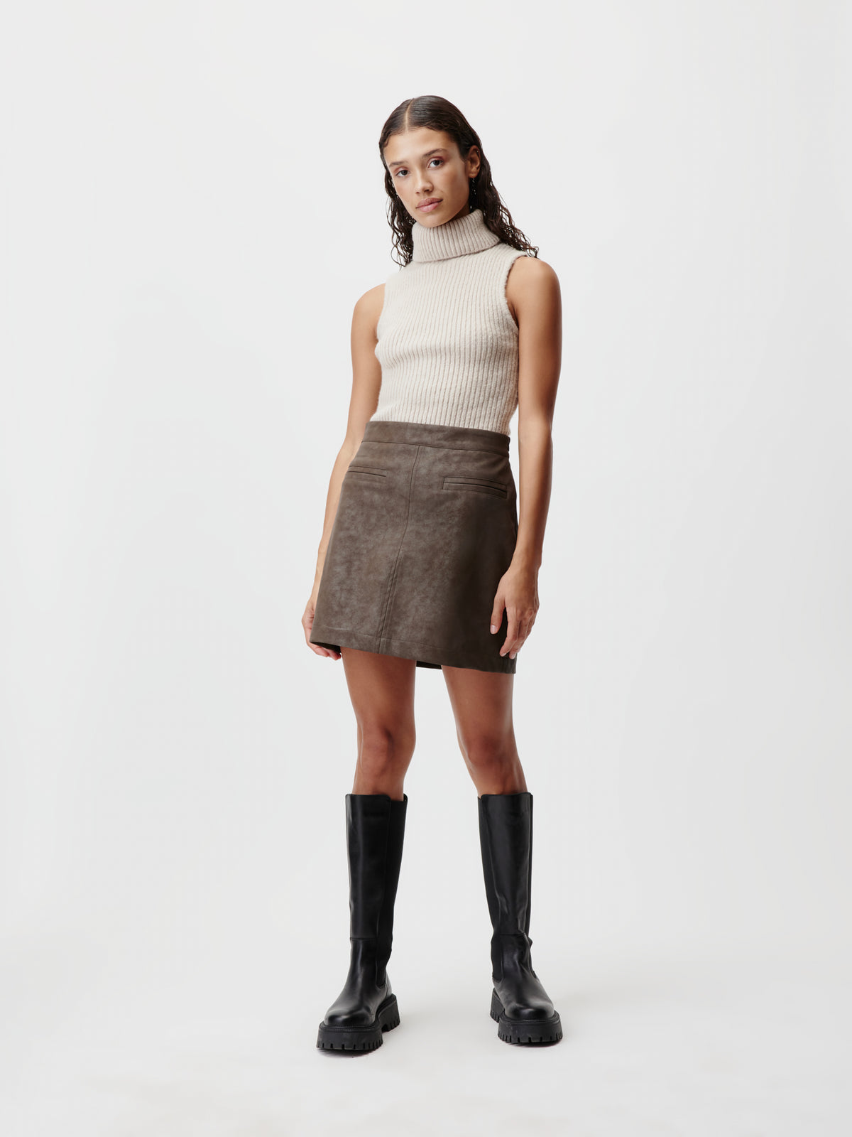 Eine Frau in einem ärmellosen, gerippten Rollkragenpullover und einem dunkelbraunen „Elisha“-Rock von LeGer by Lena Gercke, kombiniert mit schwarzen, kniehohen Stiefeln in modernem Design.