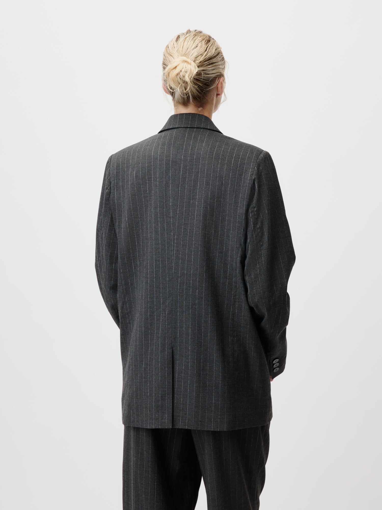 Eine Person mit blonden Haaren, die zu einem ordentlichen Dutt zusammengebunden sind, trägt das „Delphine“-Set von LeGer by Lena Gercke und präsentiert den Business-Chic, während sie mit dem Rücken zur Kamera vor einem schlichten, hellen Hintergrund steht.